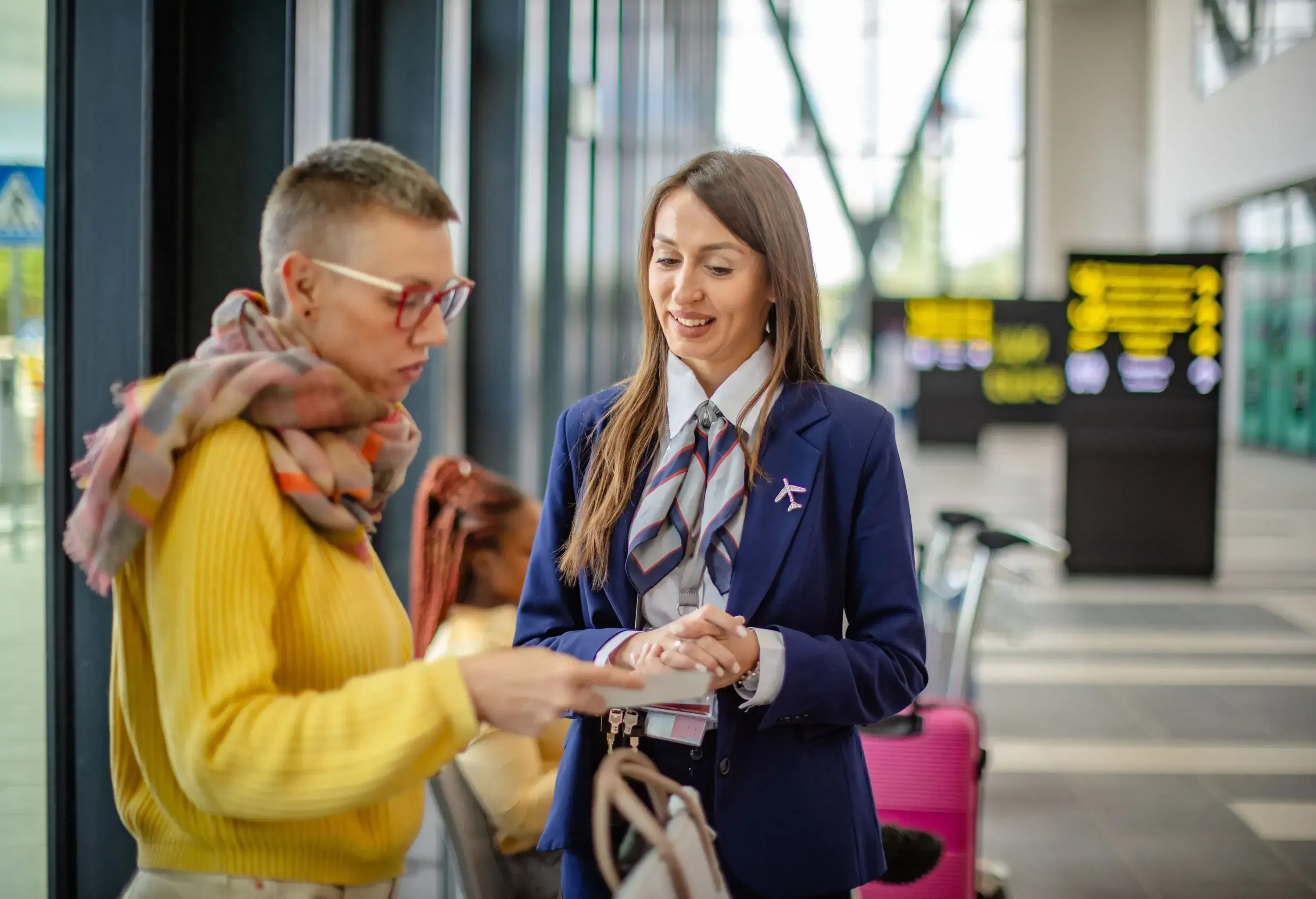 An airport staff member in a blue uniform assists a traveler dressed in yellow with travel details, set against a backdrop of a modern, busy airport terminal.
