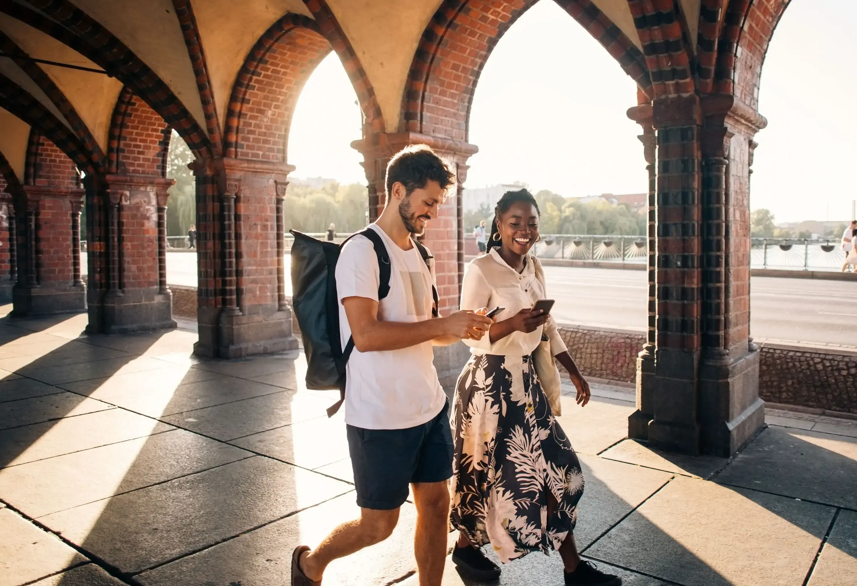 A man and a woman walk along an arched patio while navigating their smartphones.
