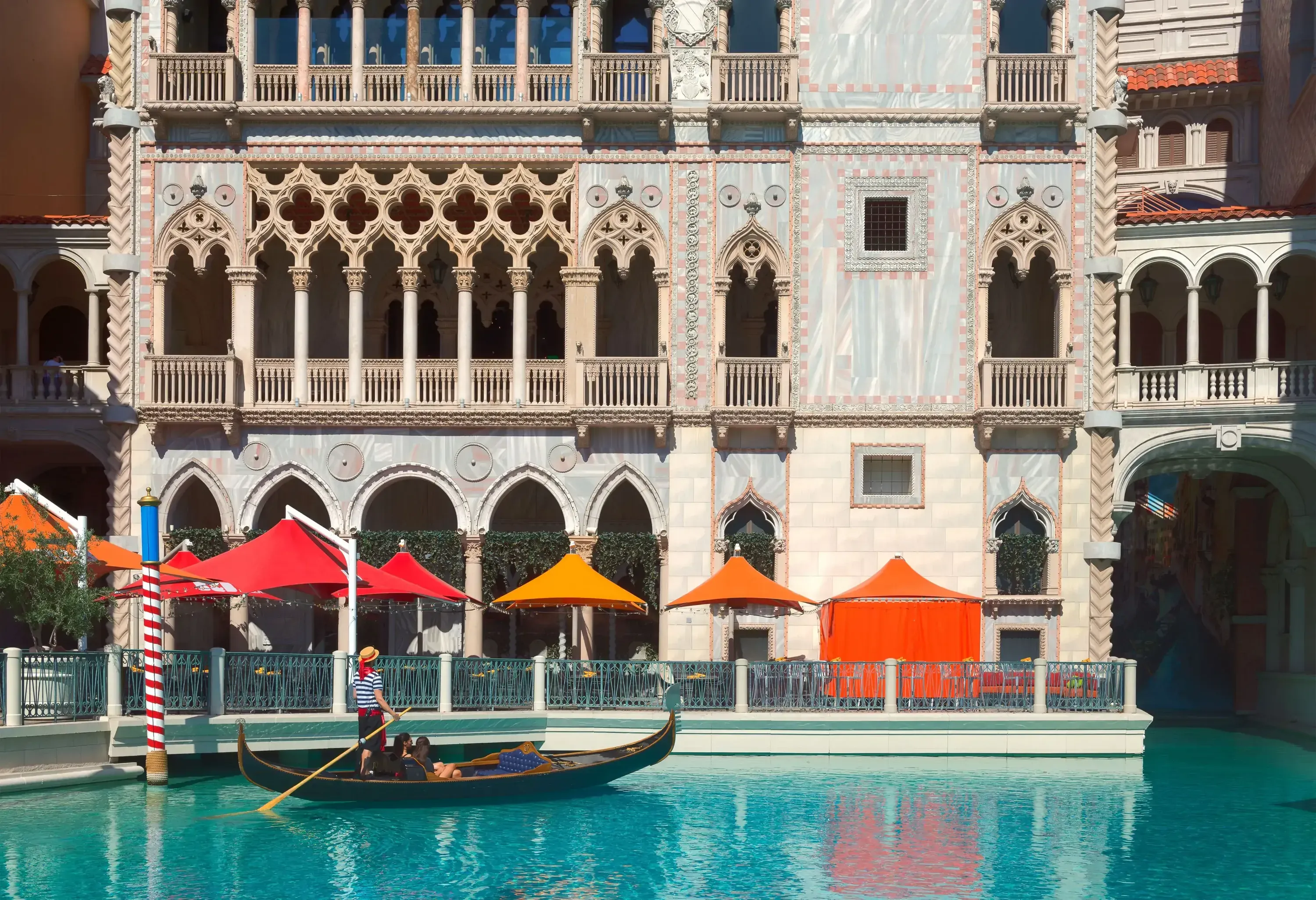 Two female friends ride on a gondola sailing through the Venetian canal along an ornate commercial building.