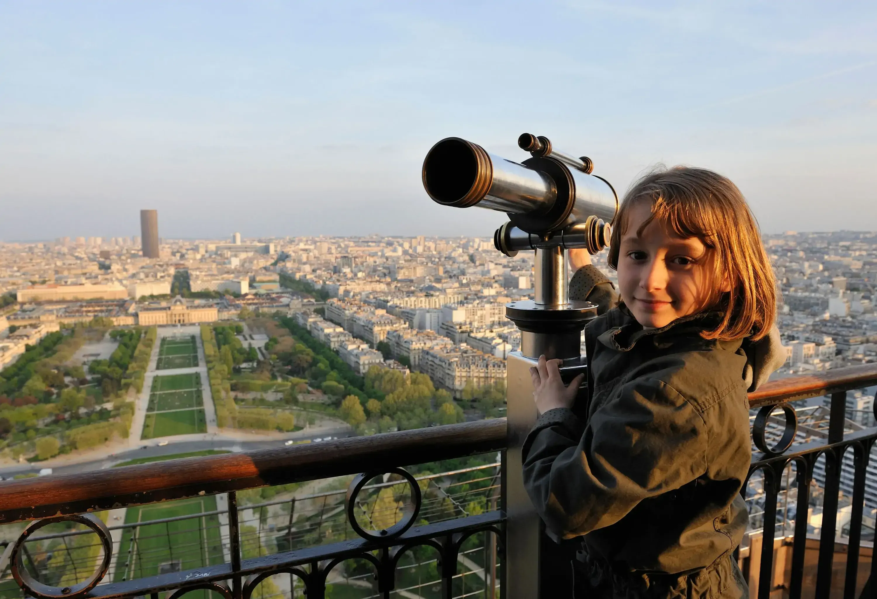 A little girl on the Eiffel tower telescope overlooking the cityscape dominated by a tower.