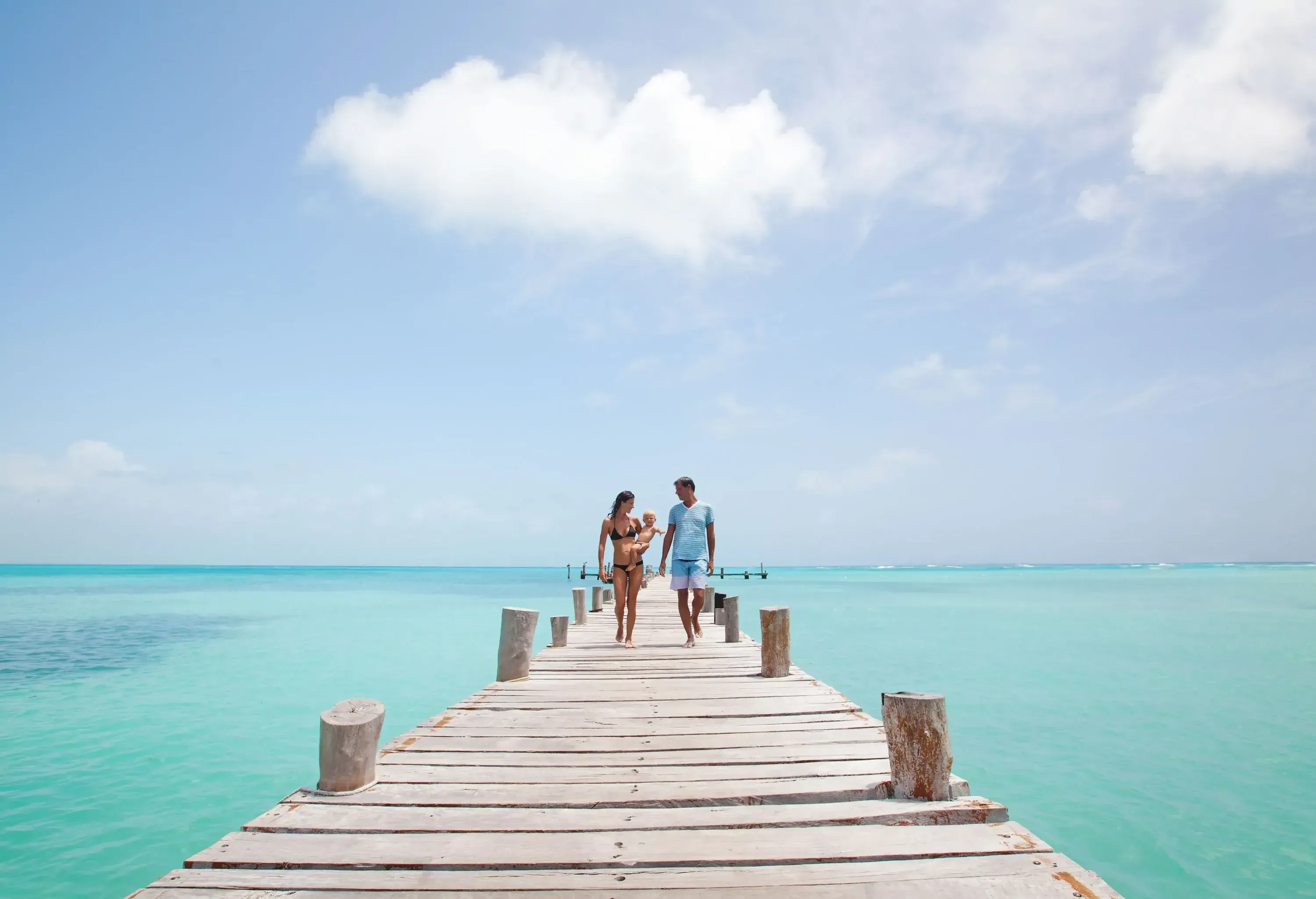 A woman in swimsuit carries a baby while walking beside a man on an overwater boardwalk.
