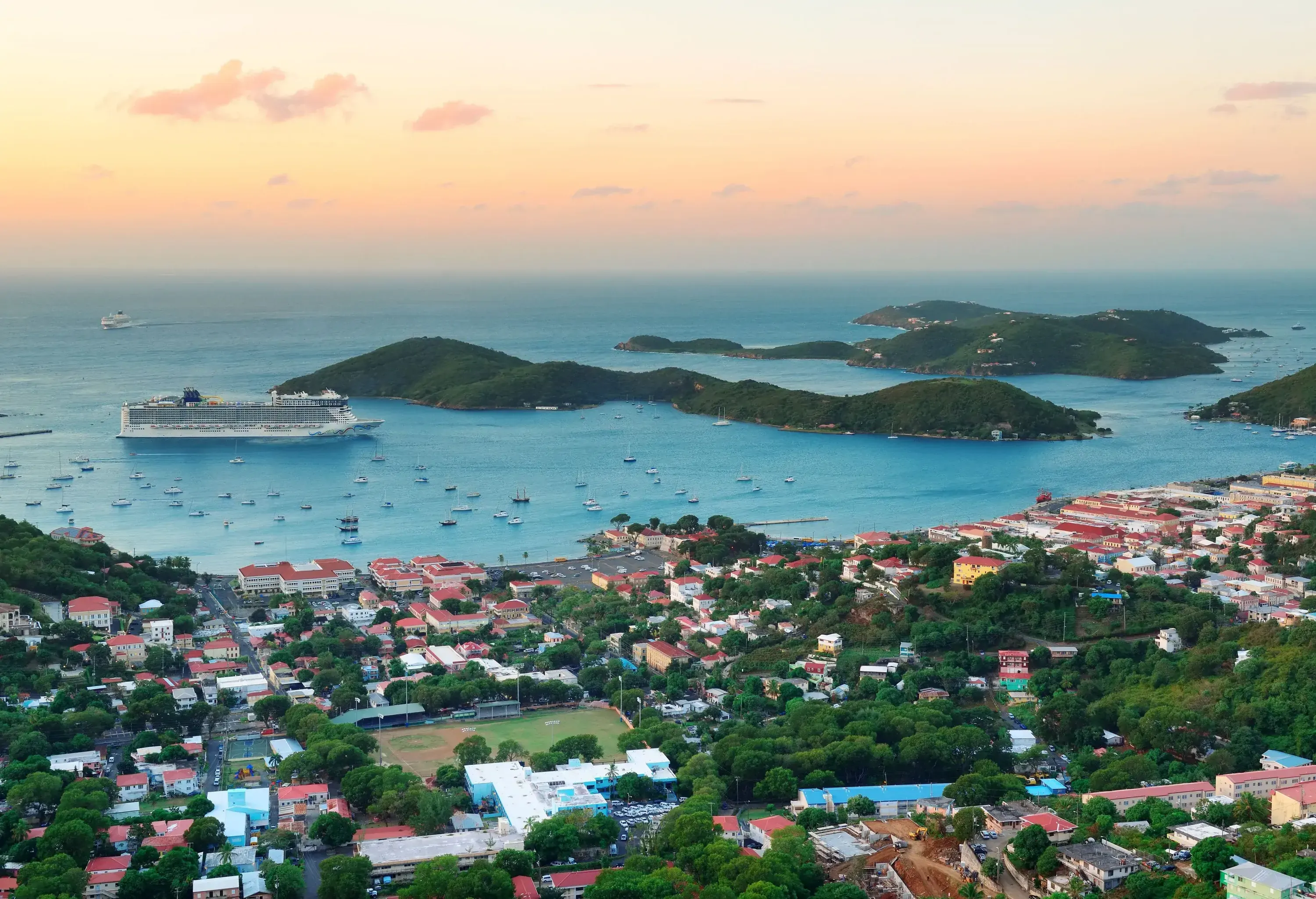A cruise ship approaches a tiny long island that emerges on the harbour with numerous small boats next to a colourful coastal city.