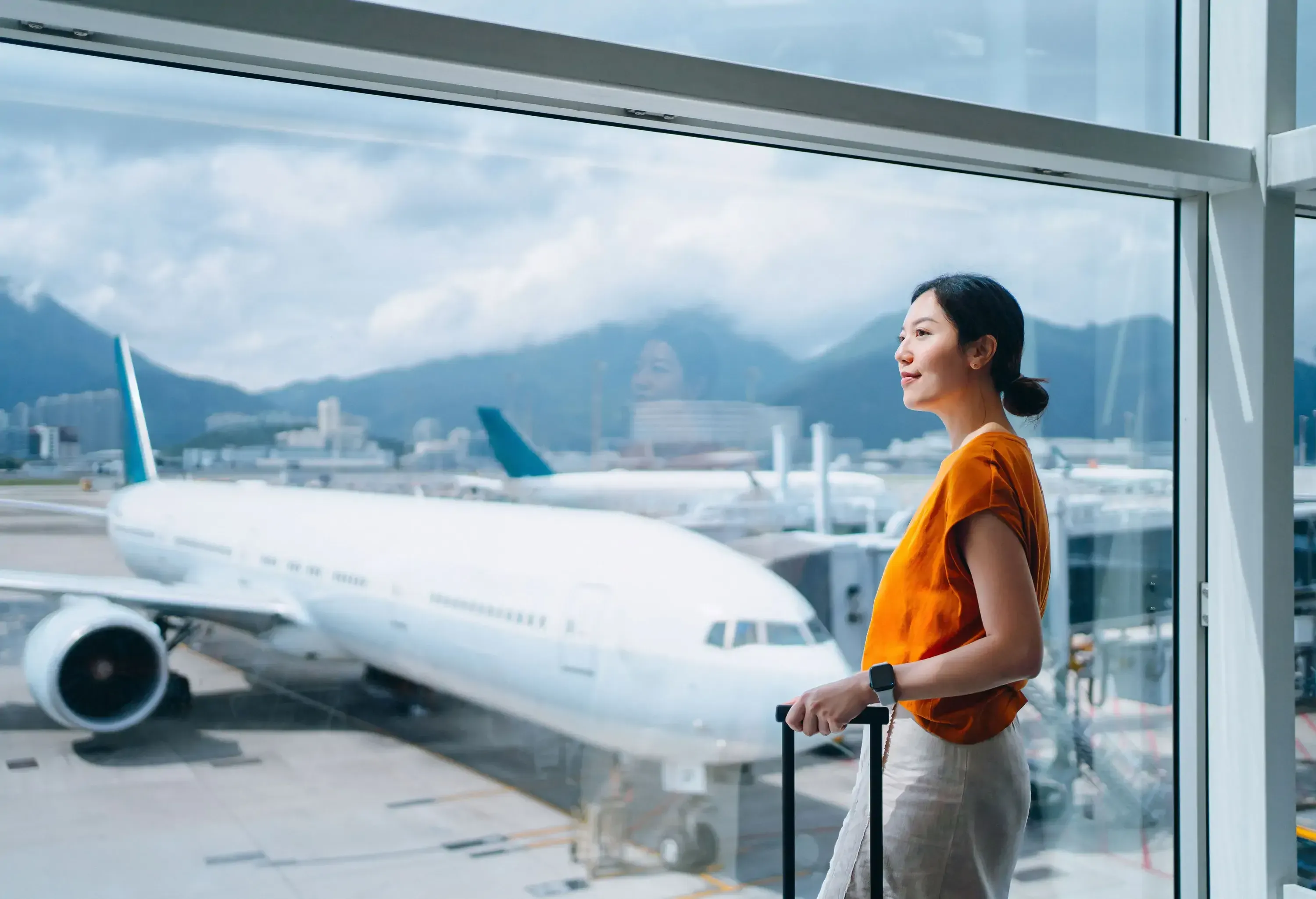 A woman carrying a suitcase looking through an airport window terminal.