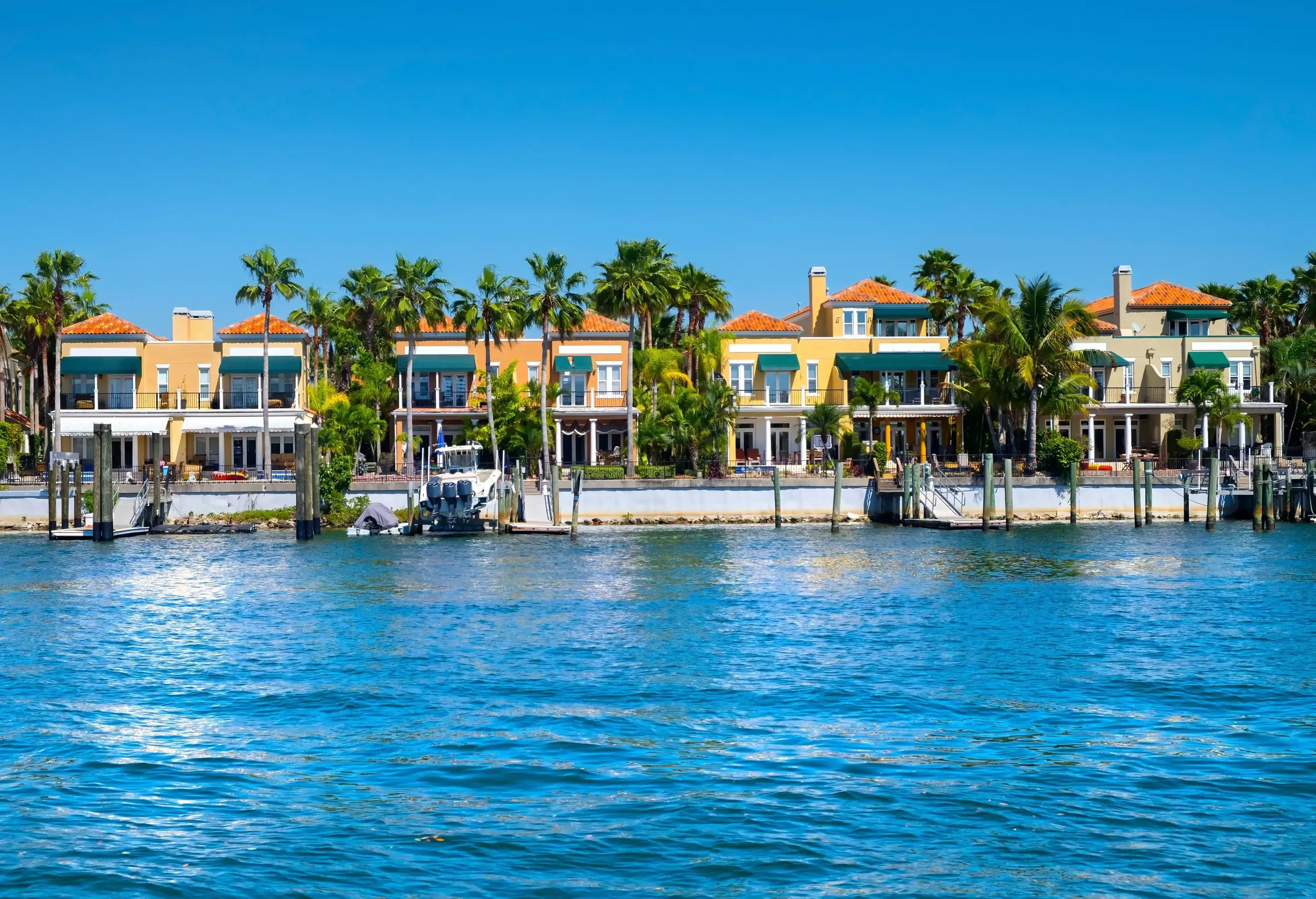 A row of large, colorful waterfront houses with docks and palm trees under a clear blue sky.