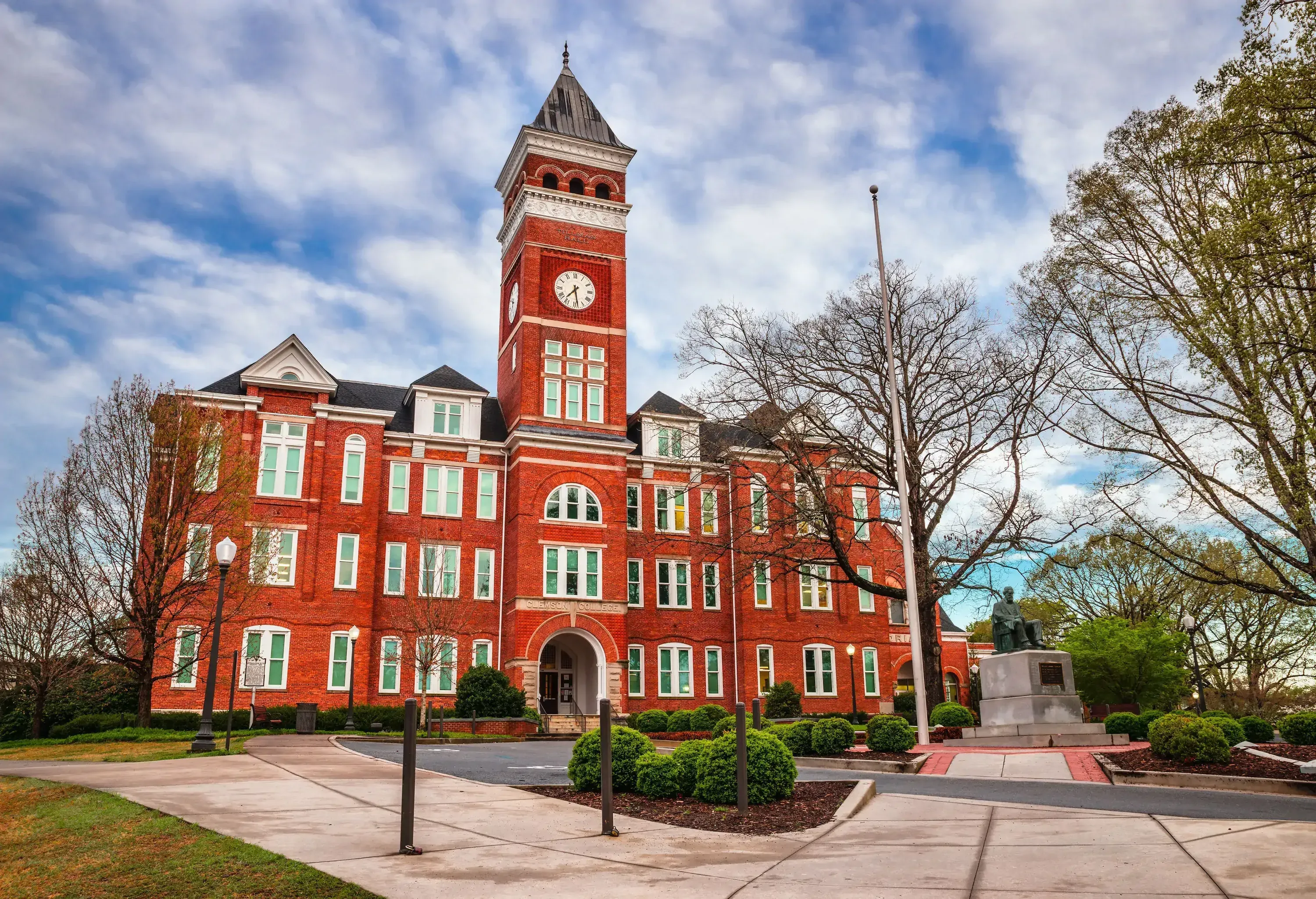 A majestic brick building stands tall, its clock tower dominating the centre, as a stoic statue stands proudly before it, surrounded by withered trees.