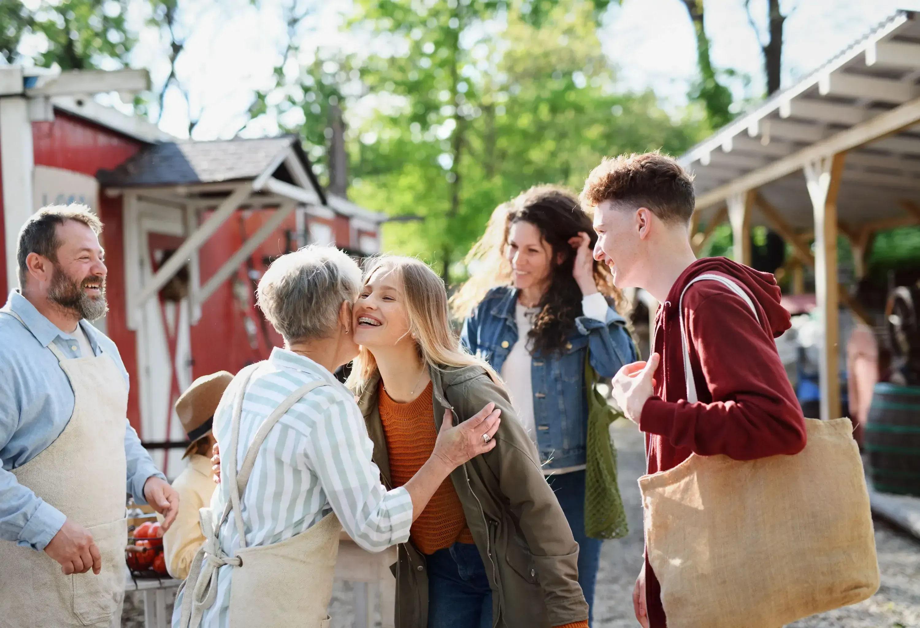 A young woman receiving a kiss on the cheek from an older woman, surrounded by smiling people at a farmer's market.