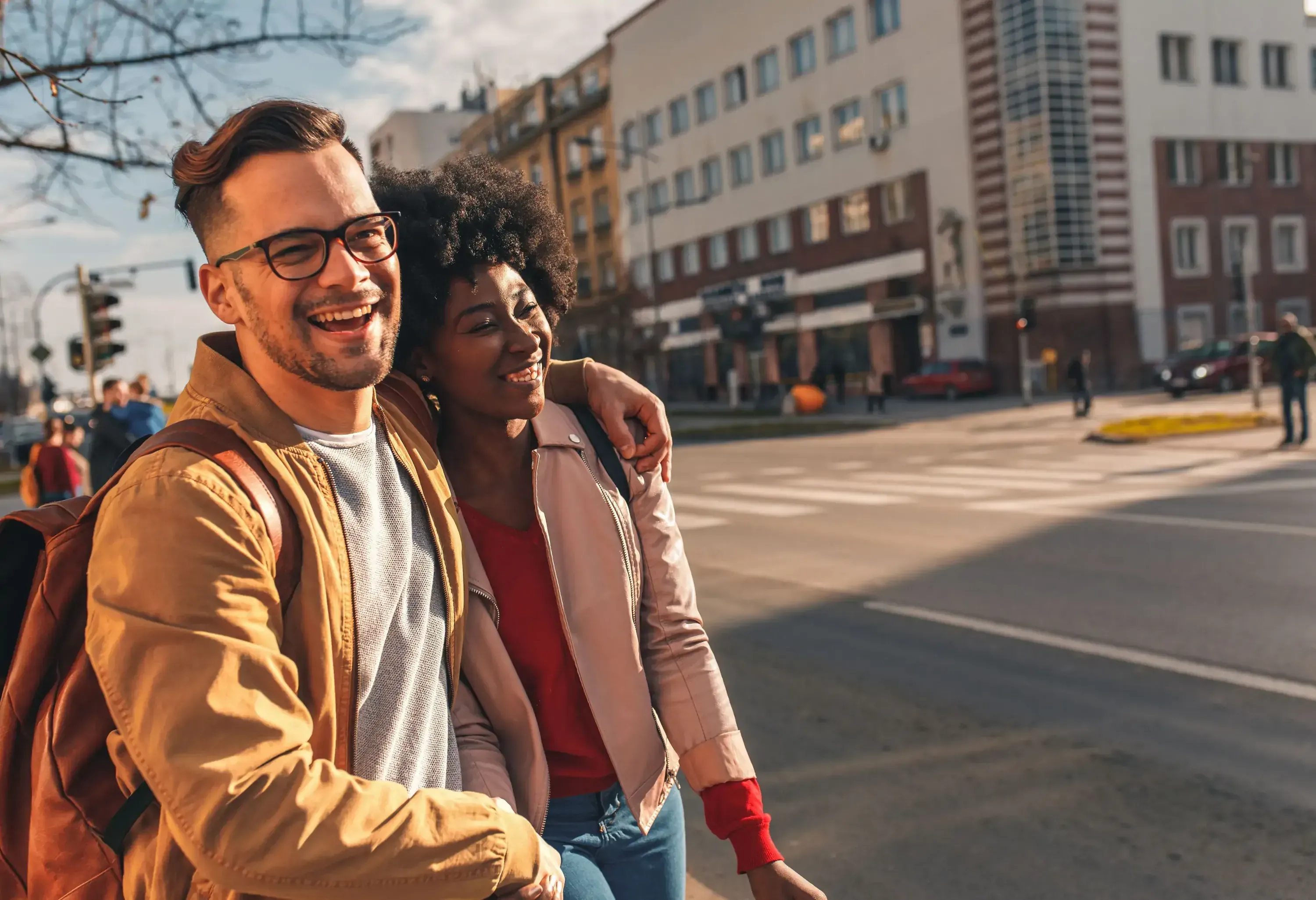 A young couple in casual clothing, holding hands and smiling while on the sidewalk.