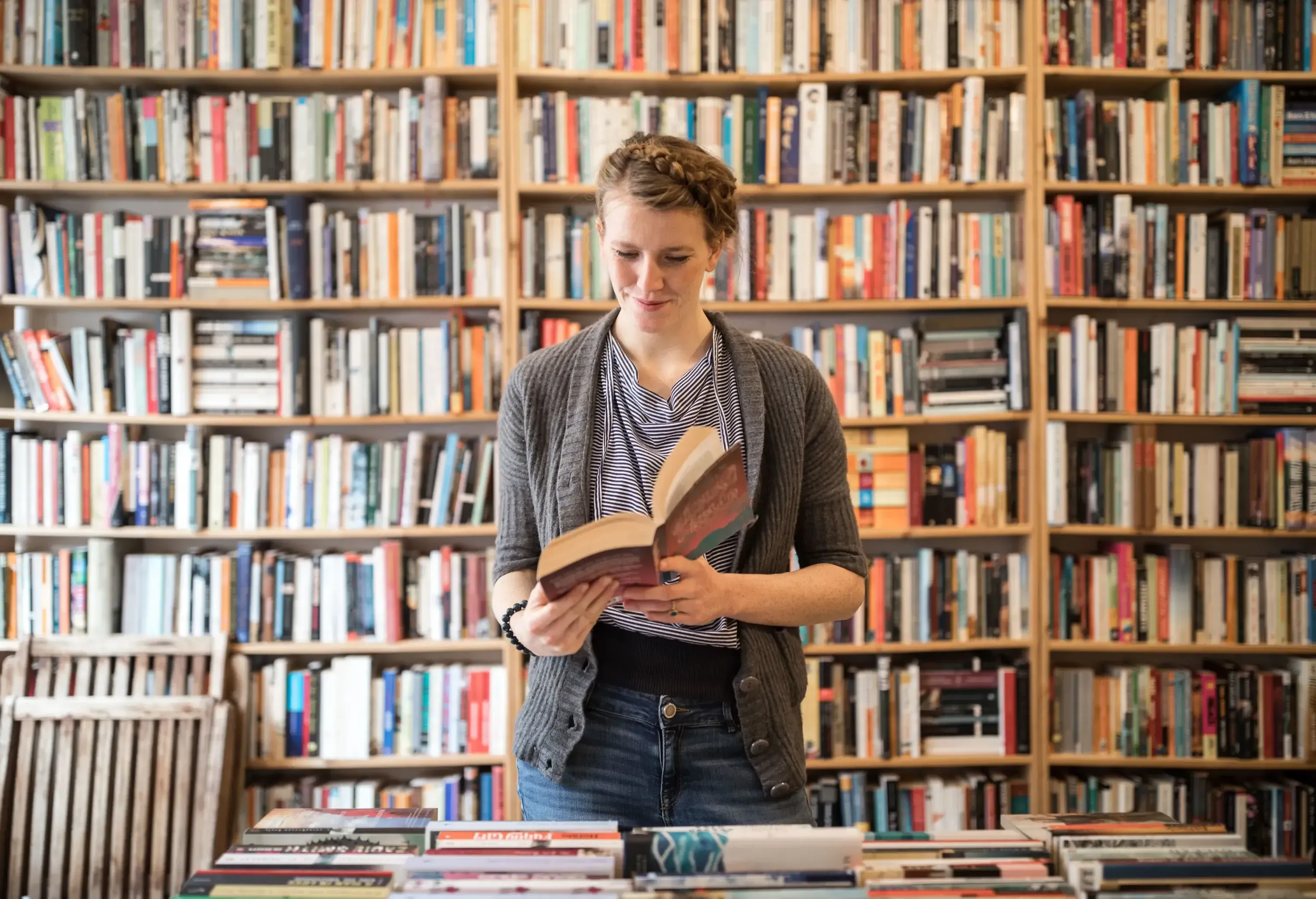 A smiling, beautiful young woman delightedly reads a book while standing against a well-stocked bookshelf in a cosy bookstore.