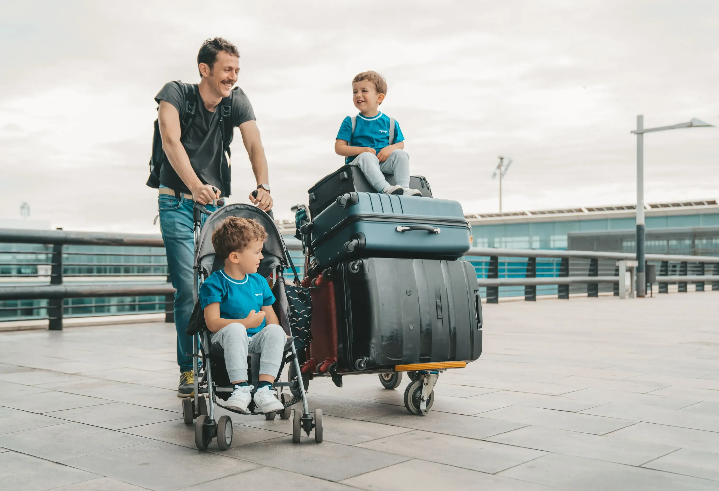 A smiling father pushes a stroller with his young son while his other son sits on a tall stack of luggage next to him.