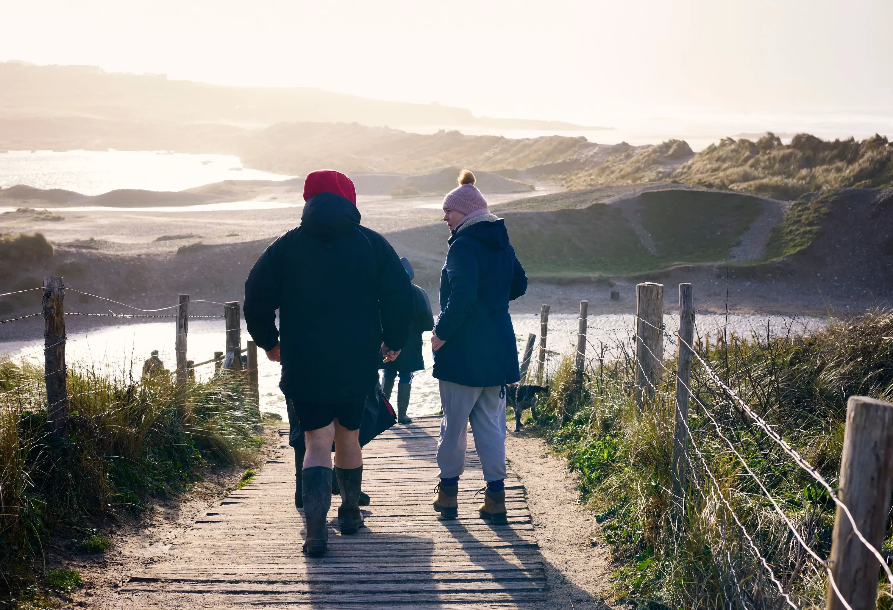 A group of people walking on a wooden path towards a body of water bordered by sand dunes under hazy skies.