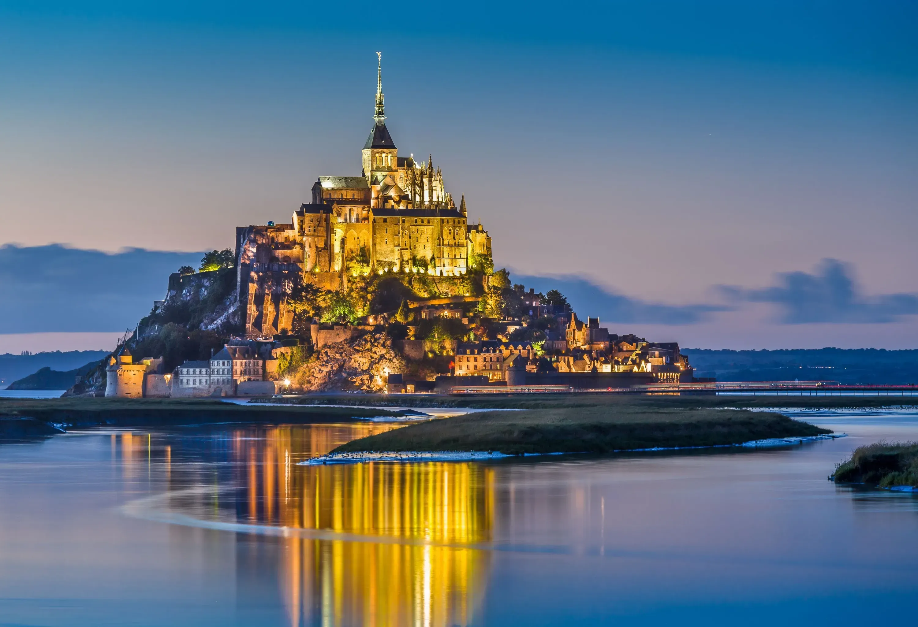 A night-time scene of an illuminated town perched on a hill of an island with a towering spire on top.