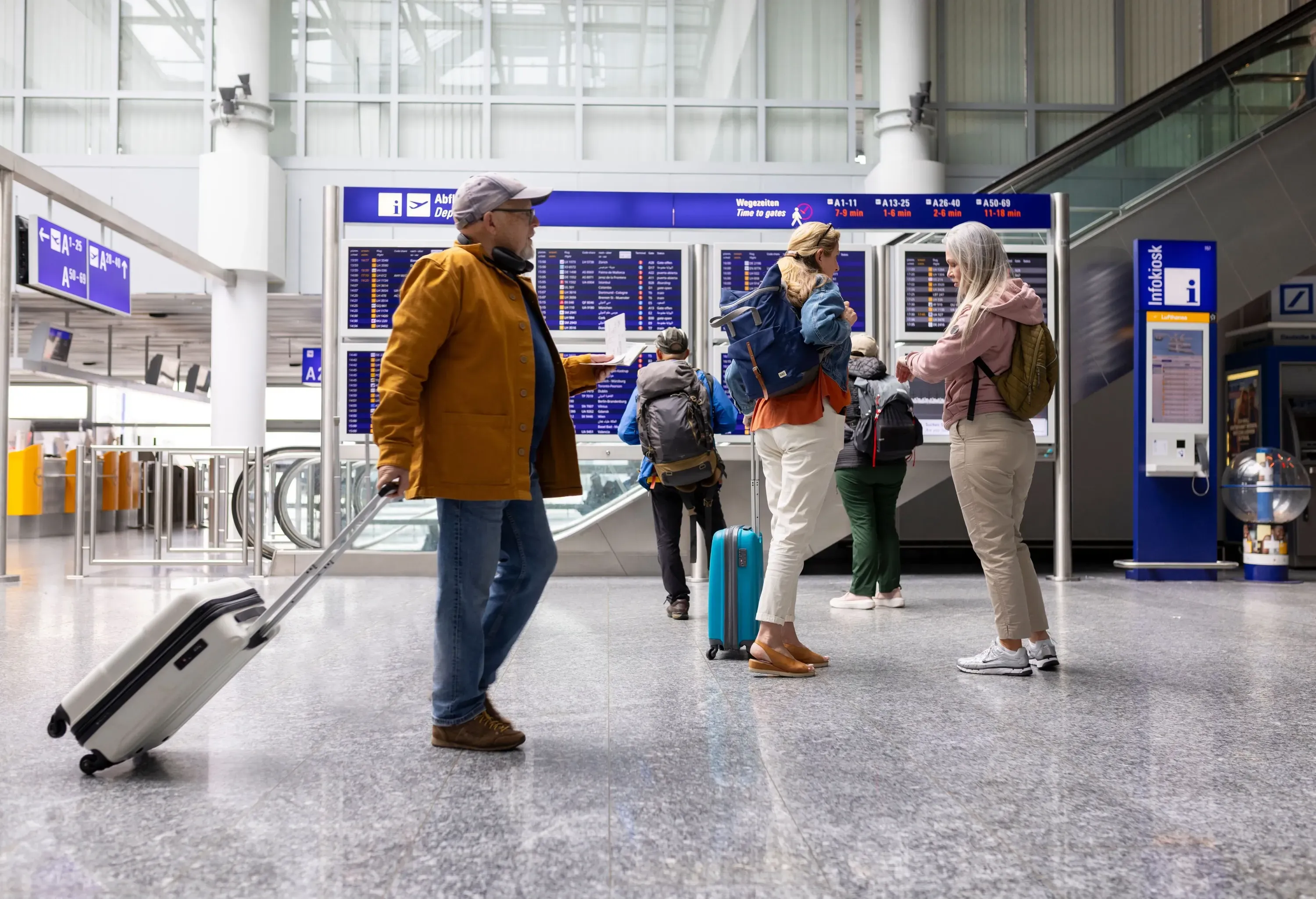 Wide shot of travellers standing and walking around the airport in front of the departure boards. Two women are standing together and talking to each other. A man is walking past them with a suitcase while holding his plane ticket.