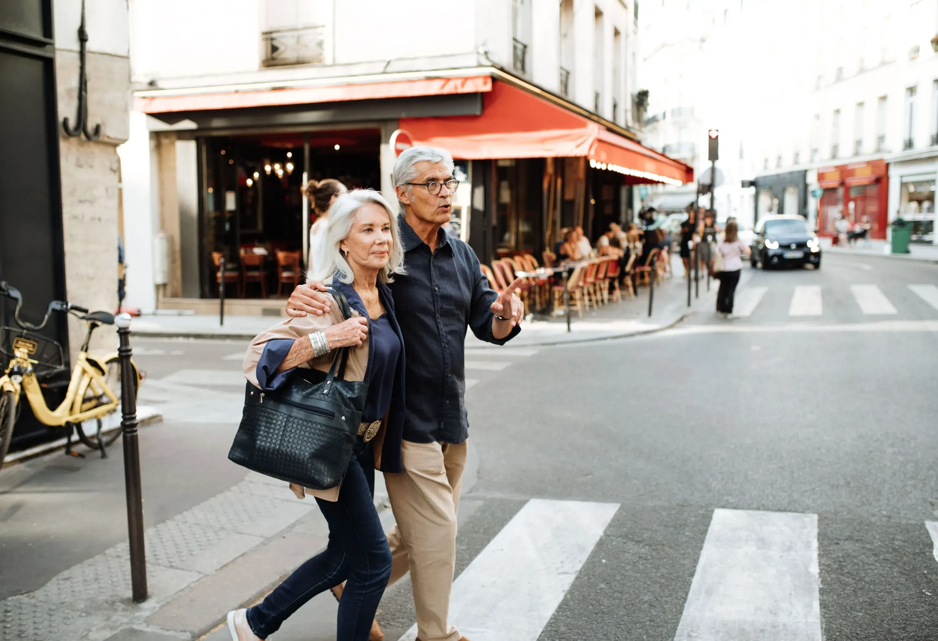 A lovely senior couple gracefully crosses the pedestrian lane, with a charming caf&eacute; building standing proudly in the background.
