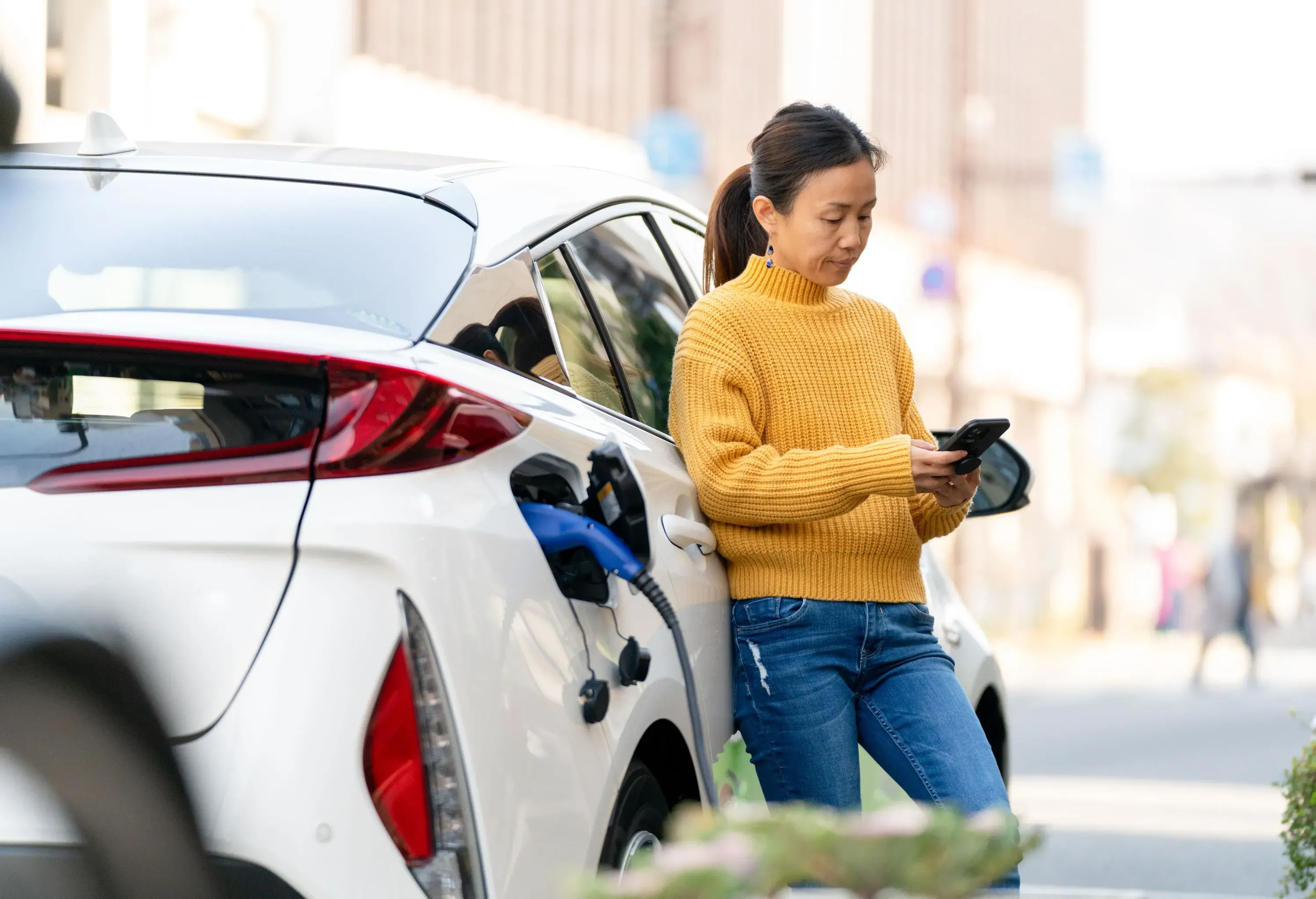 A woman looks at her mobile phone while charging her white electric car.