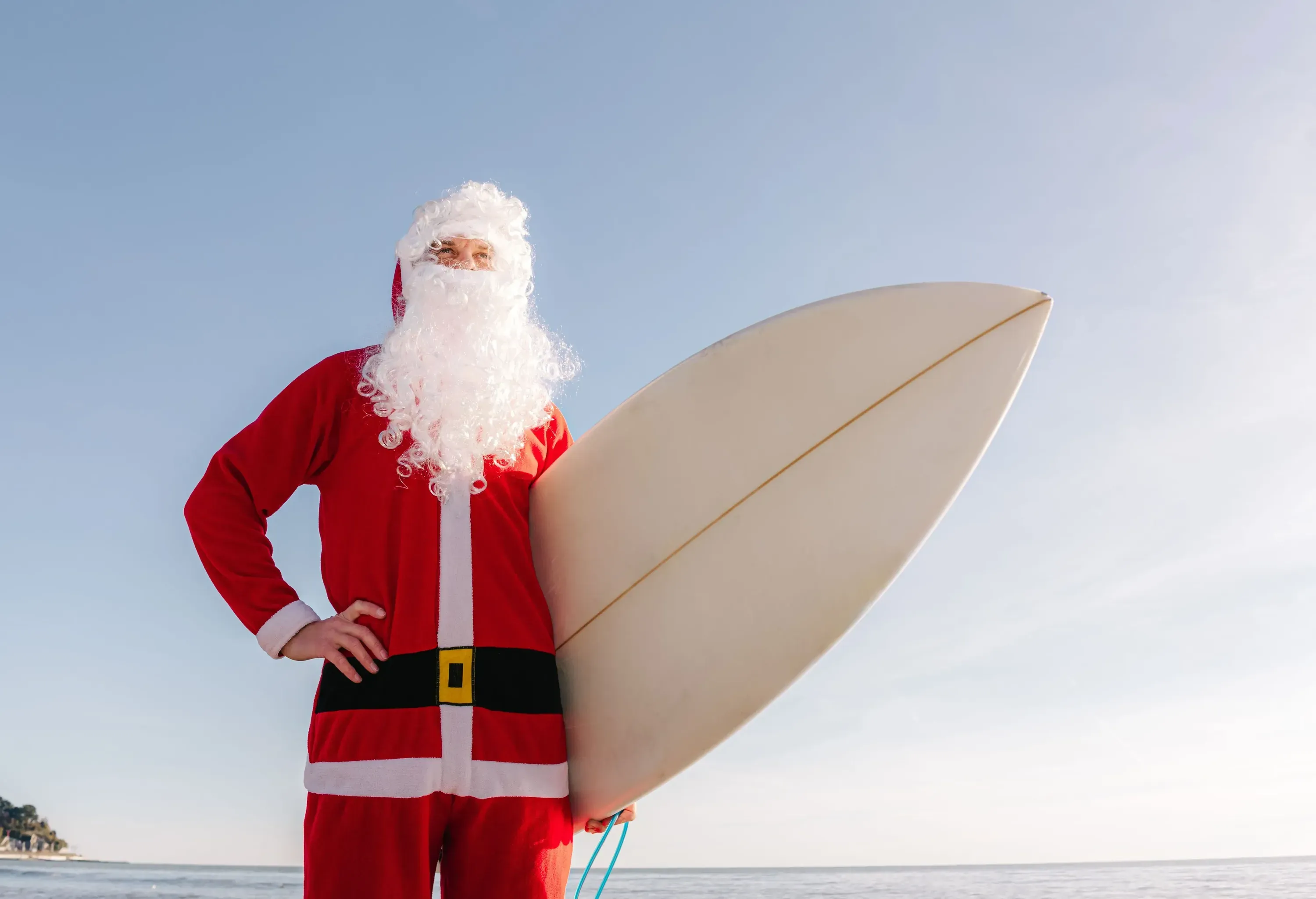 A person dressed as Santa Claus stands on a beach, holding a surfboard under a clear sky.
