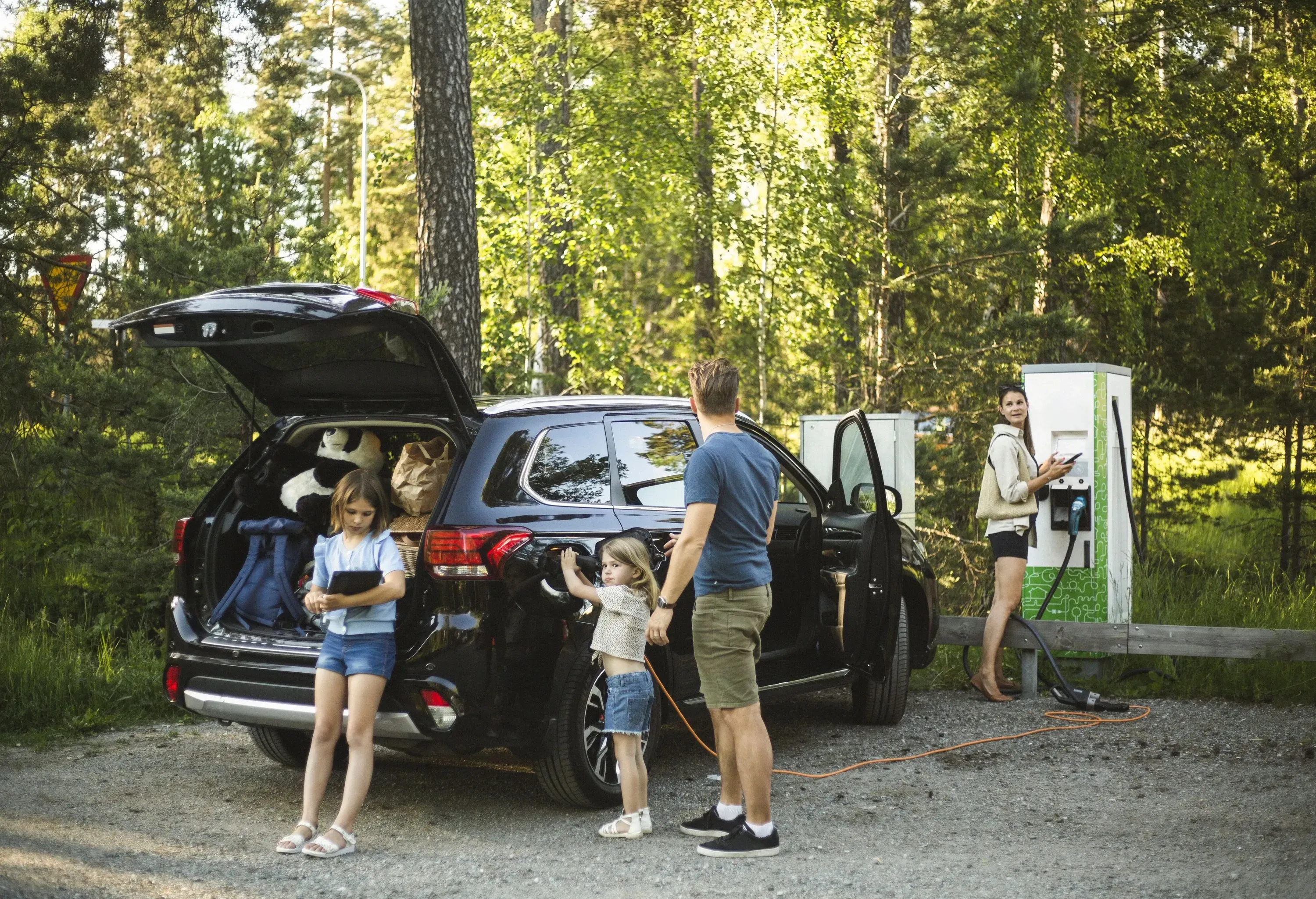 A family of four hanging outside as they charge their electric car in a forested area.