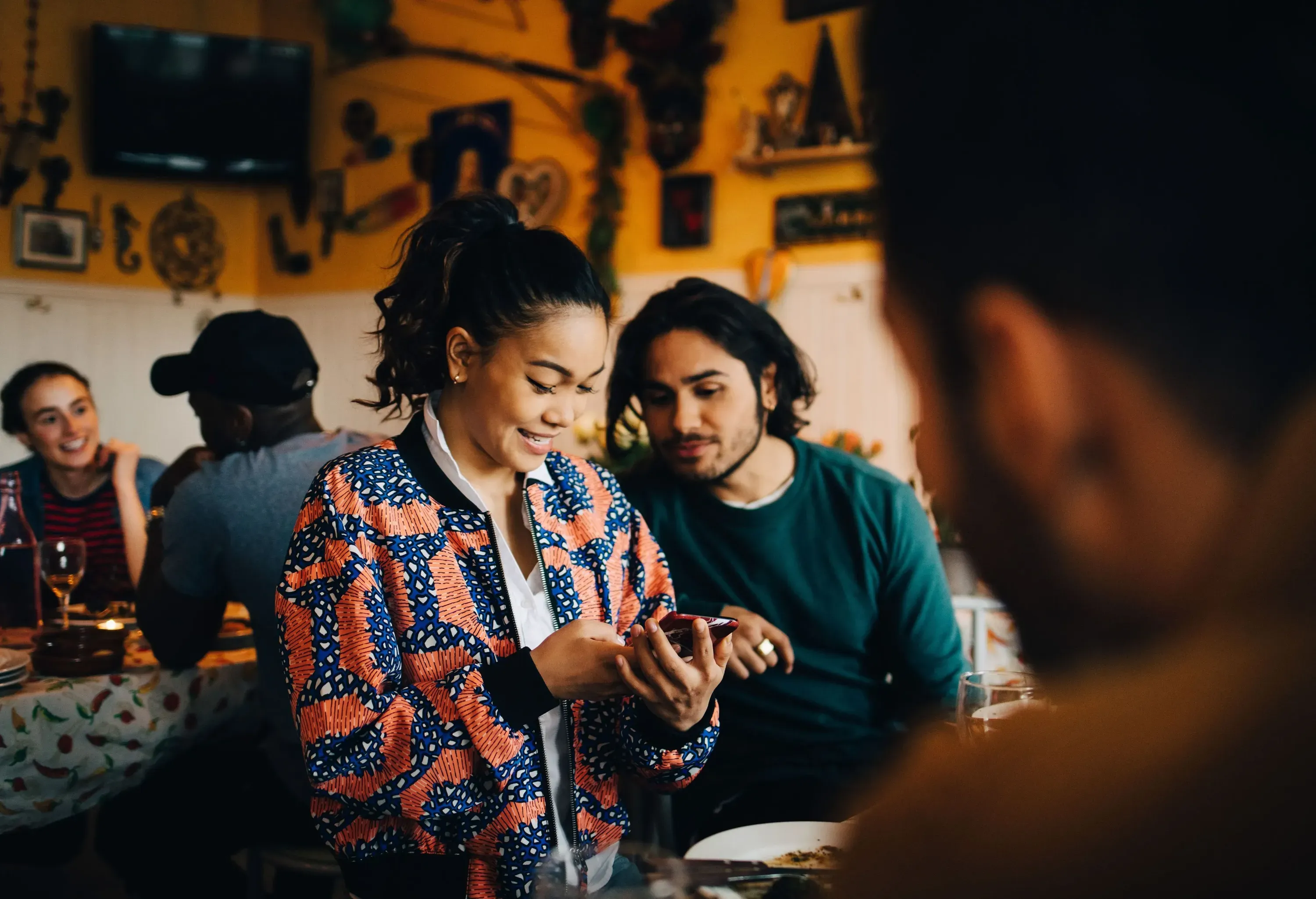 A woman in a colourful jacket smiles and looks at her smartphone while a male friend observes.