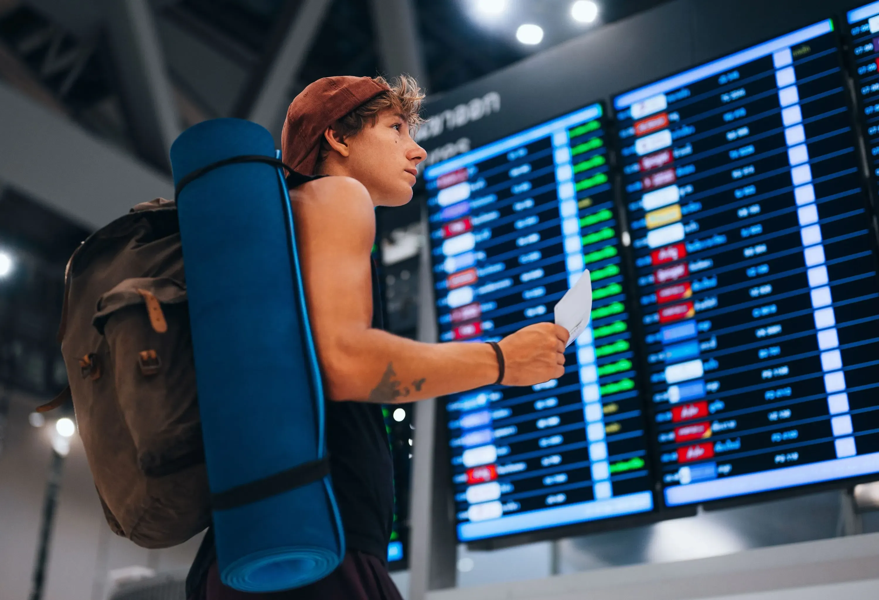 A man with a large backpack and a rolled blue sleeping mat looks up at an airport departures board, holding a boarding pass.