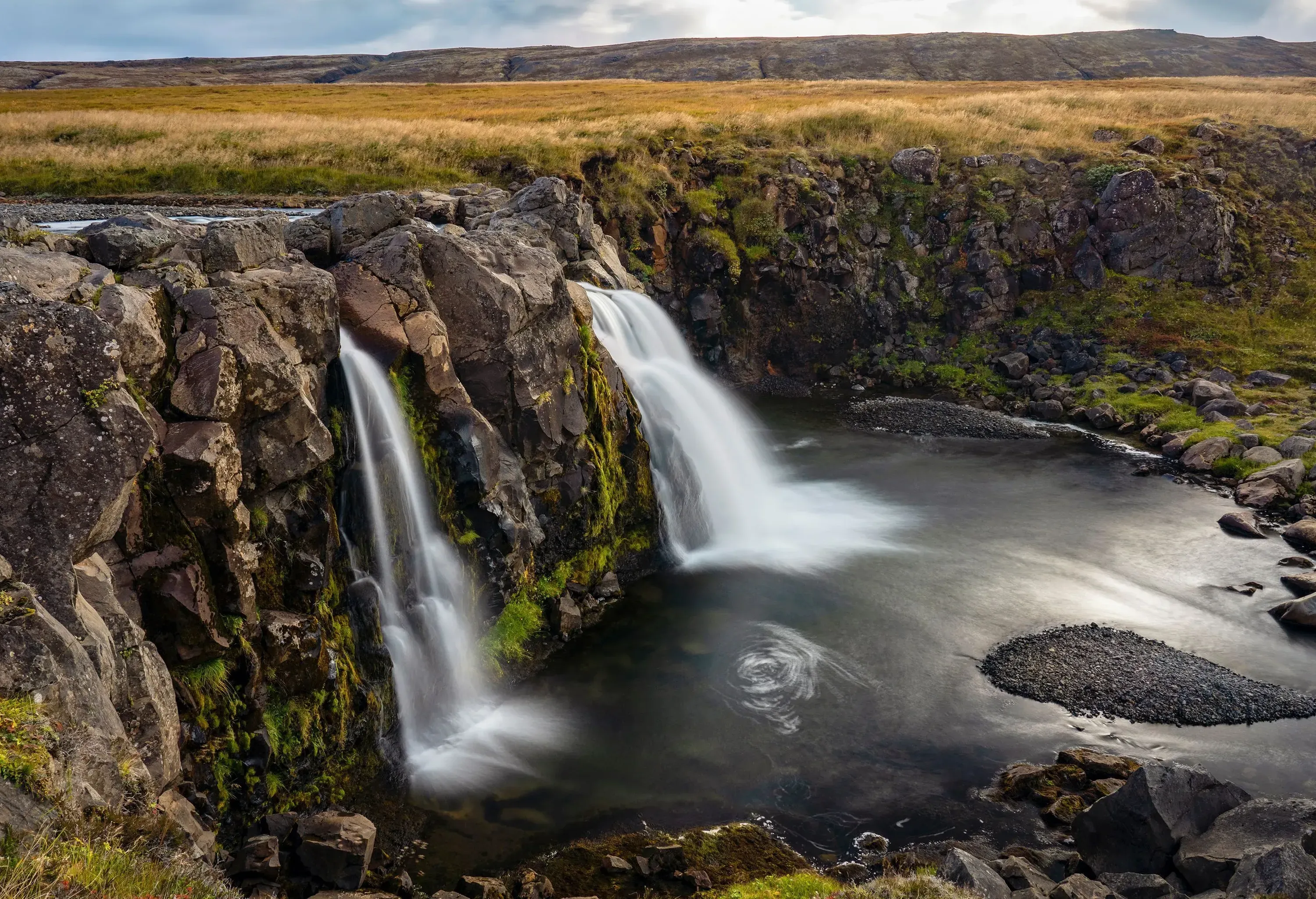Two beautiful waterfalls cascade down a steep cliff into a crystal-clear pool below, surrounded by lush natural surroundings and grassy ground.
