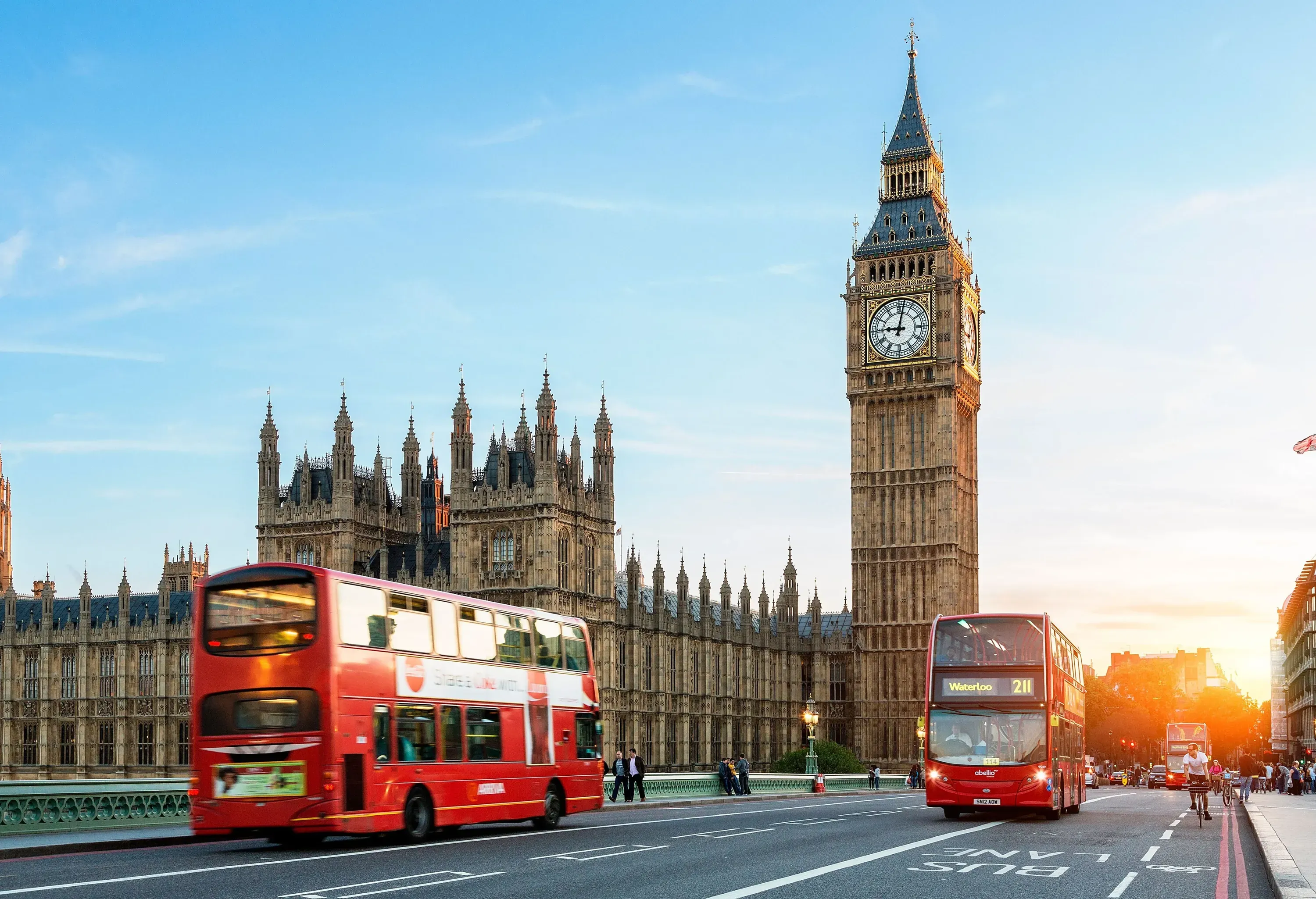 Two red double-decker buses on a sunlit street with a large clock tower and a grand building in the background.