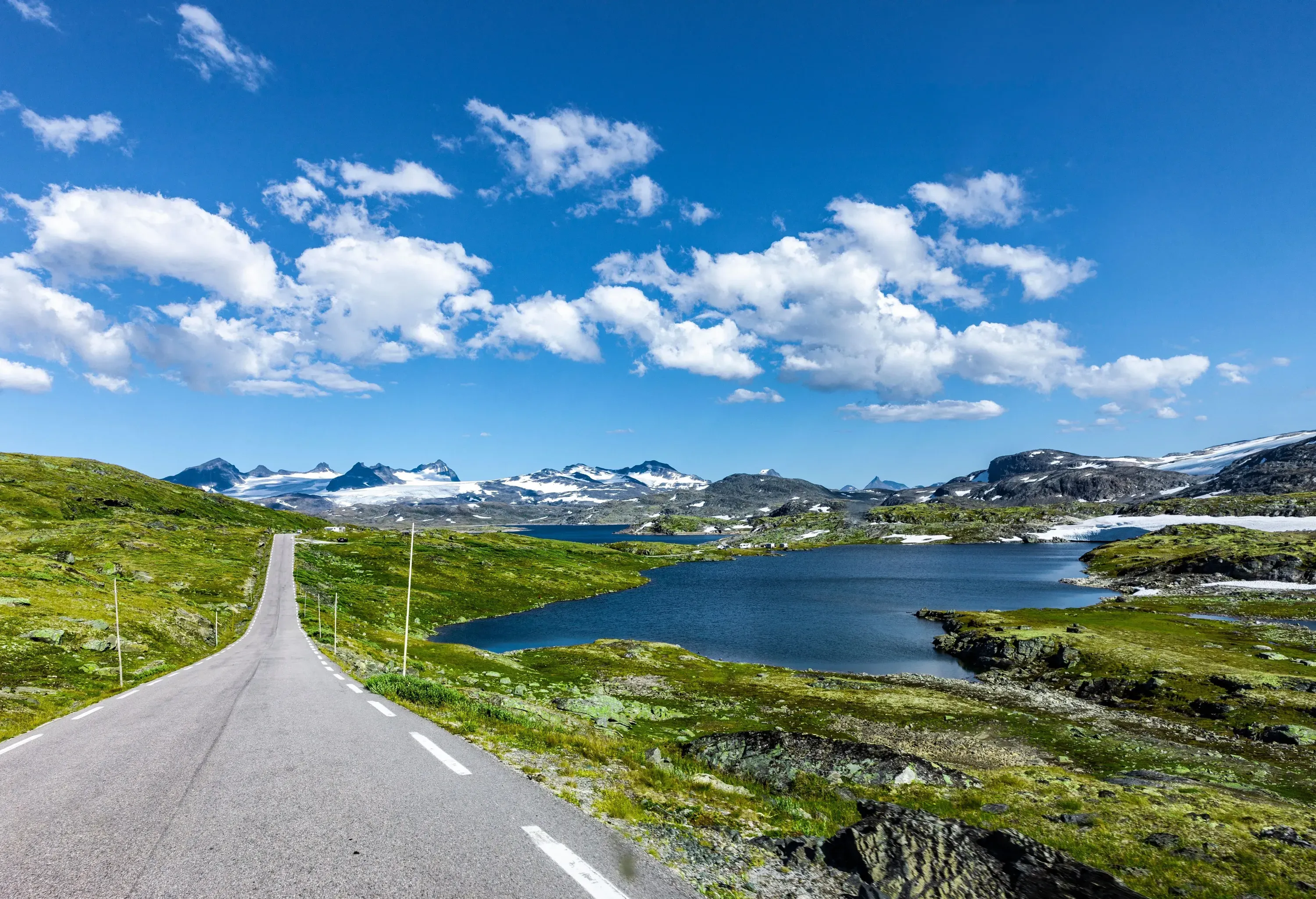 An empty road with views of a lake and snow-capped mountains.