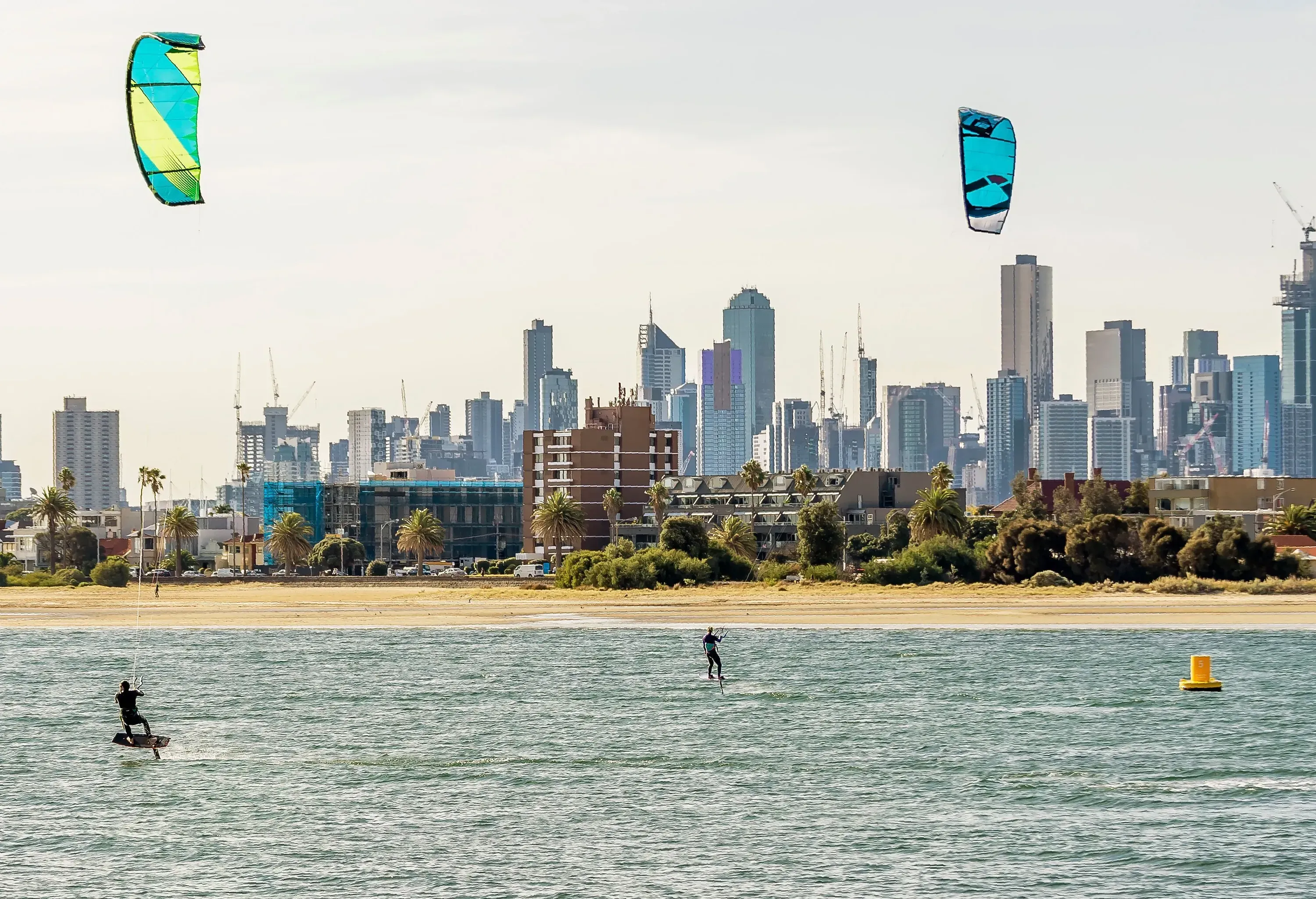 On a cloudy day, two male friends are kiteboarding on the bay against the city skyline.