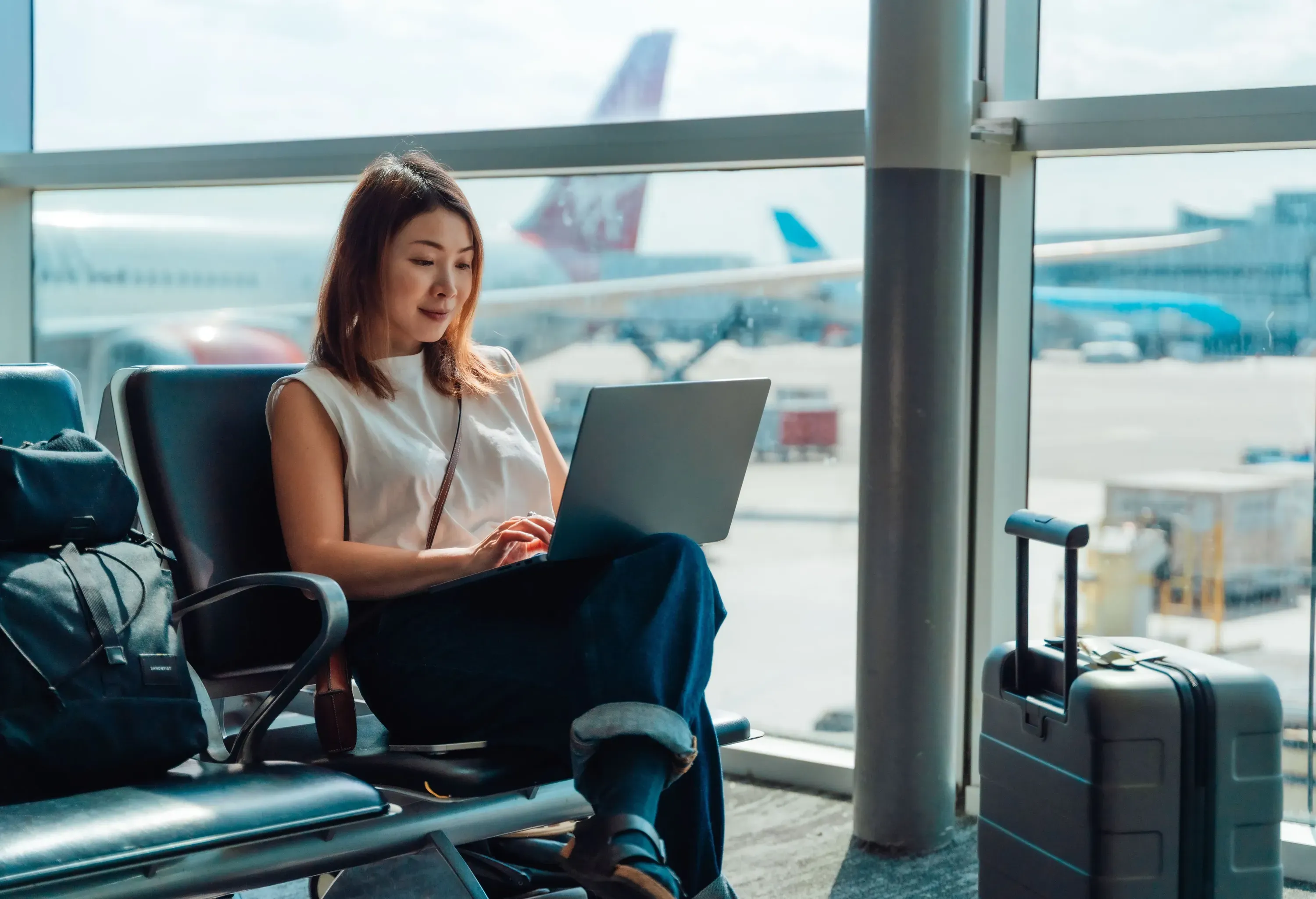 Young Asian woman checking emails, tracking flight status, booking car rental, making a hotel reservations with laptop, sitting at airport terminal before departure. Business travel and vacations.