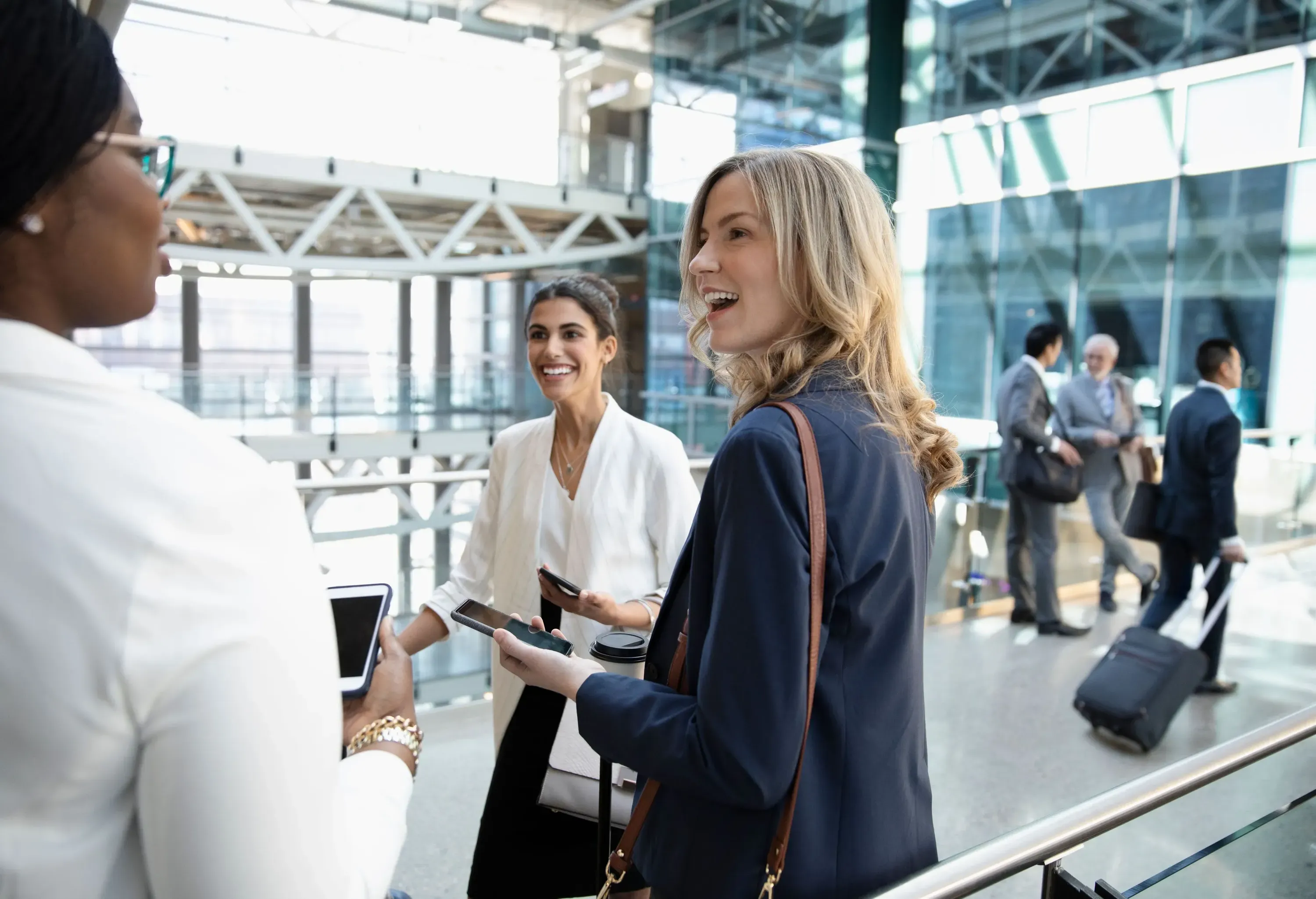 A group of female employees standing and conversing together while holding their phones, waiting for their flight at the airport.