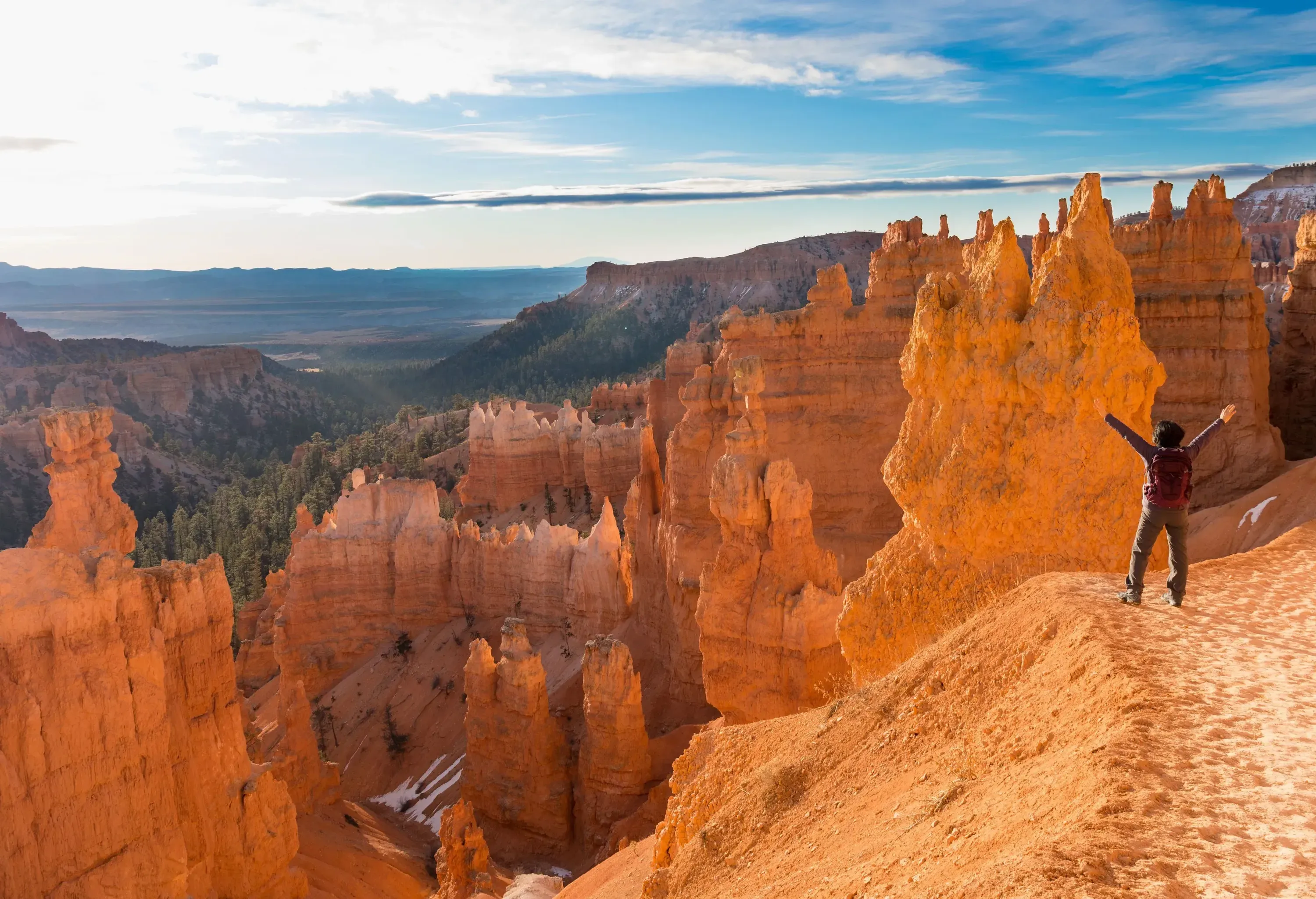 A hiker with their arms lifted in the air, standing atop a hill surrounded by hoodoos.