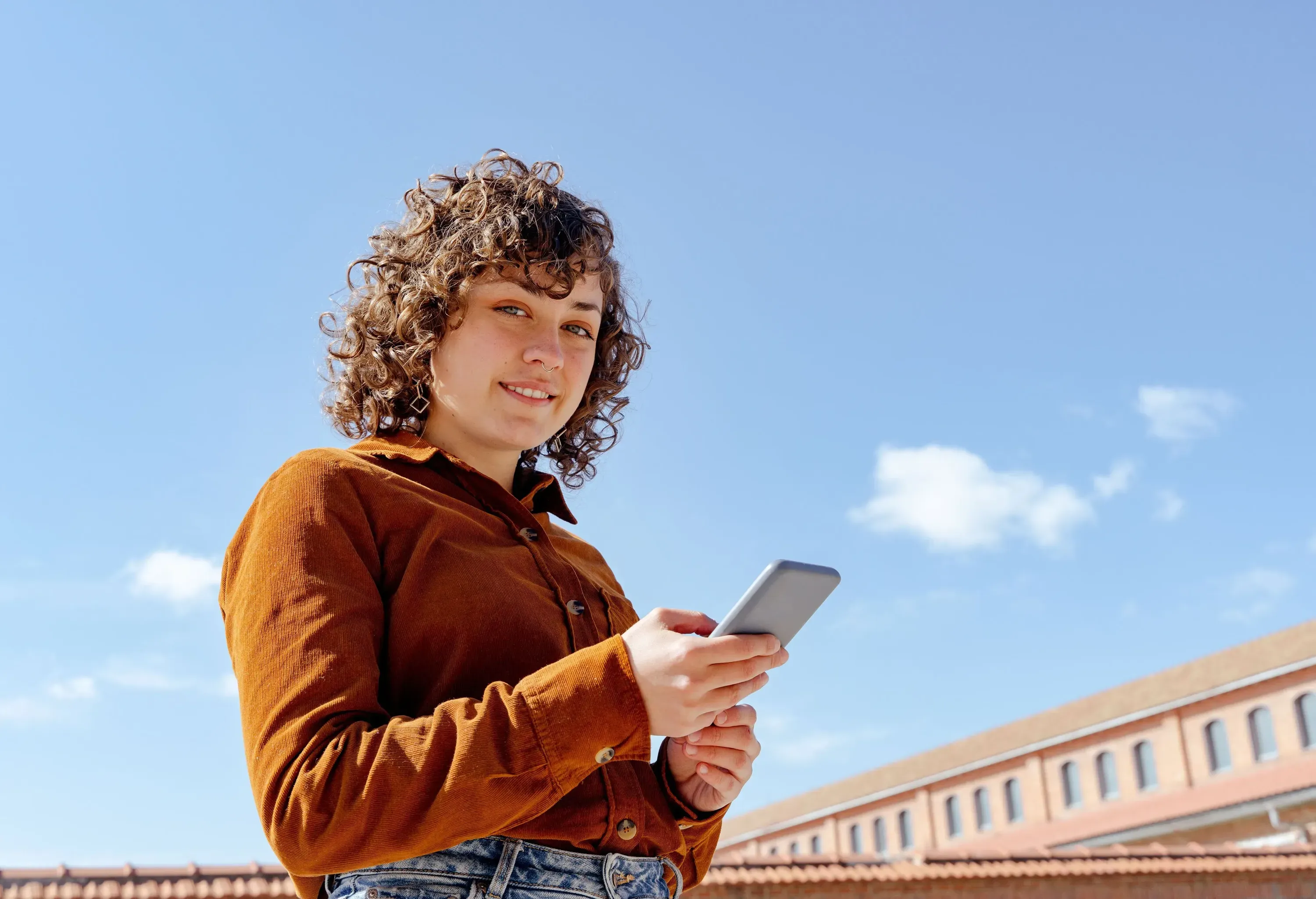A smiling young woman in a brown corduroy shirt holding a smartphone outdoors under a clear blue sky.