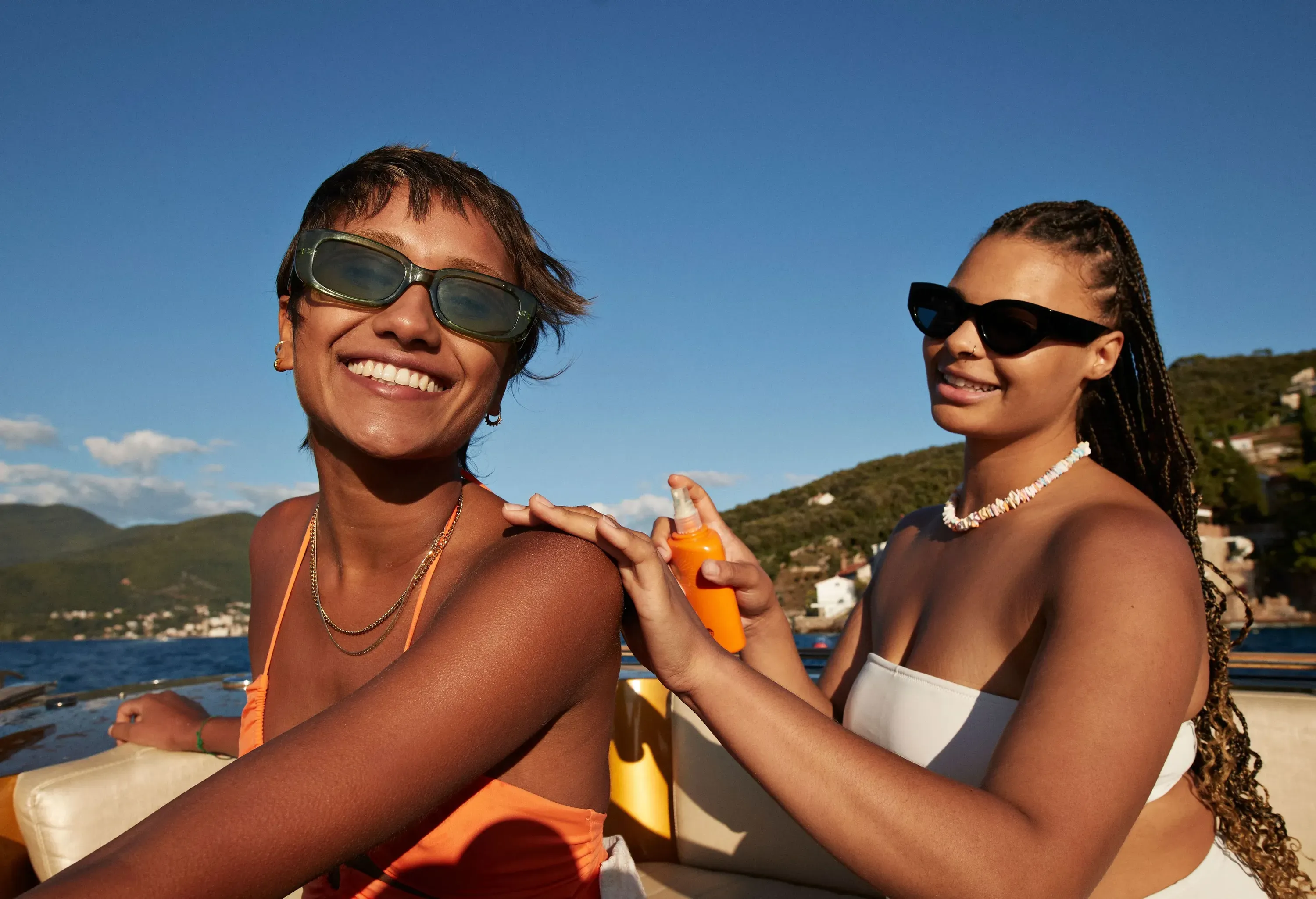 One individual applies sunscreen to another person’s shoulder in a boat on calm water surrounded by mountains under clear skies.