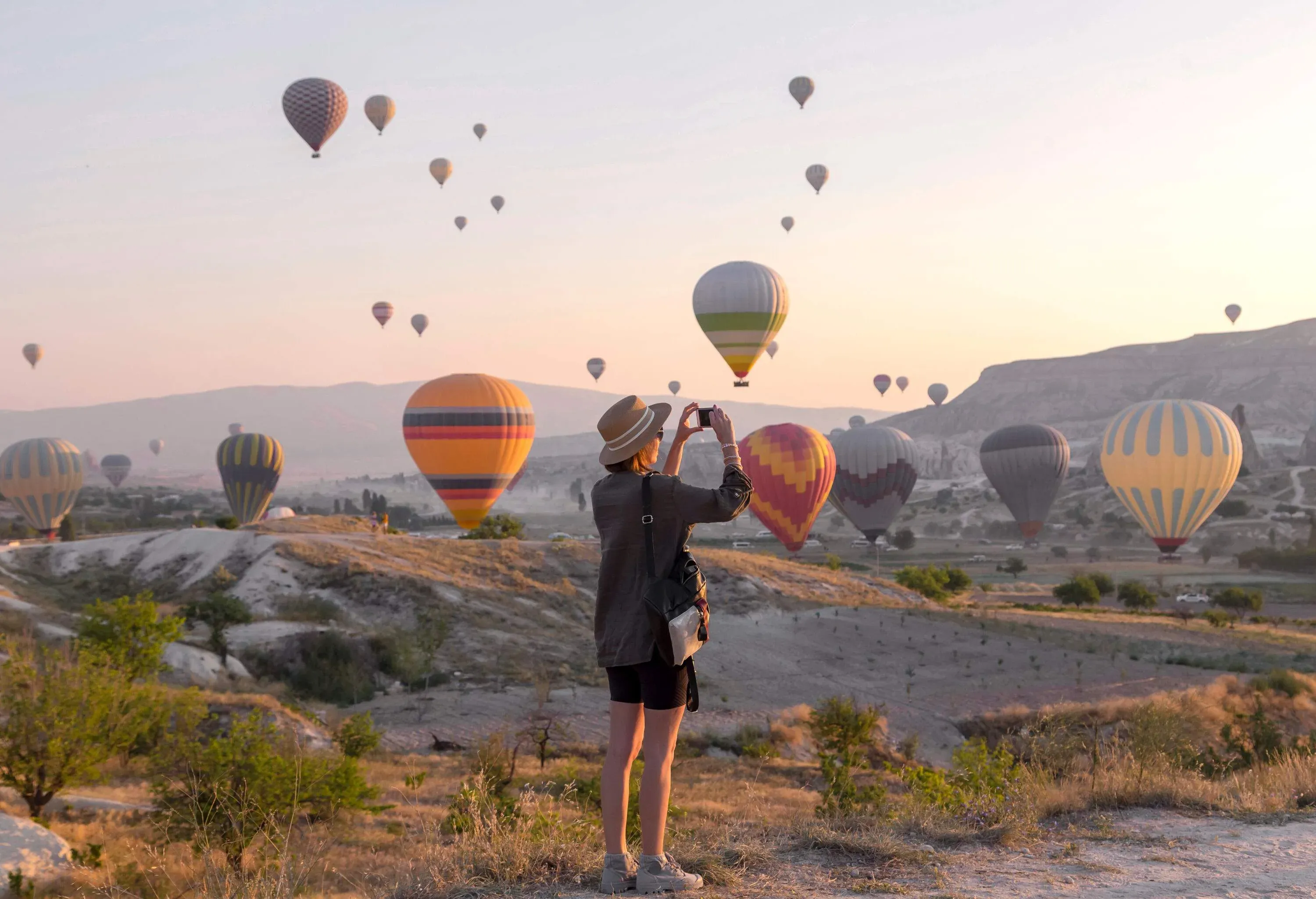 A tourist taking a photo of the hot air balloons hovering barely above the ground and soaring in the sky.
