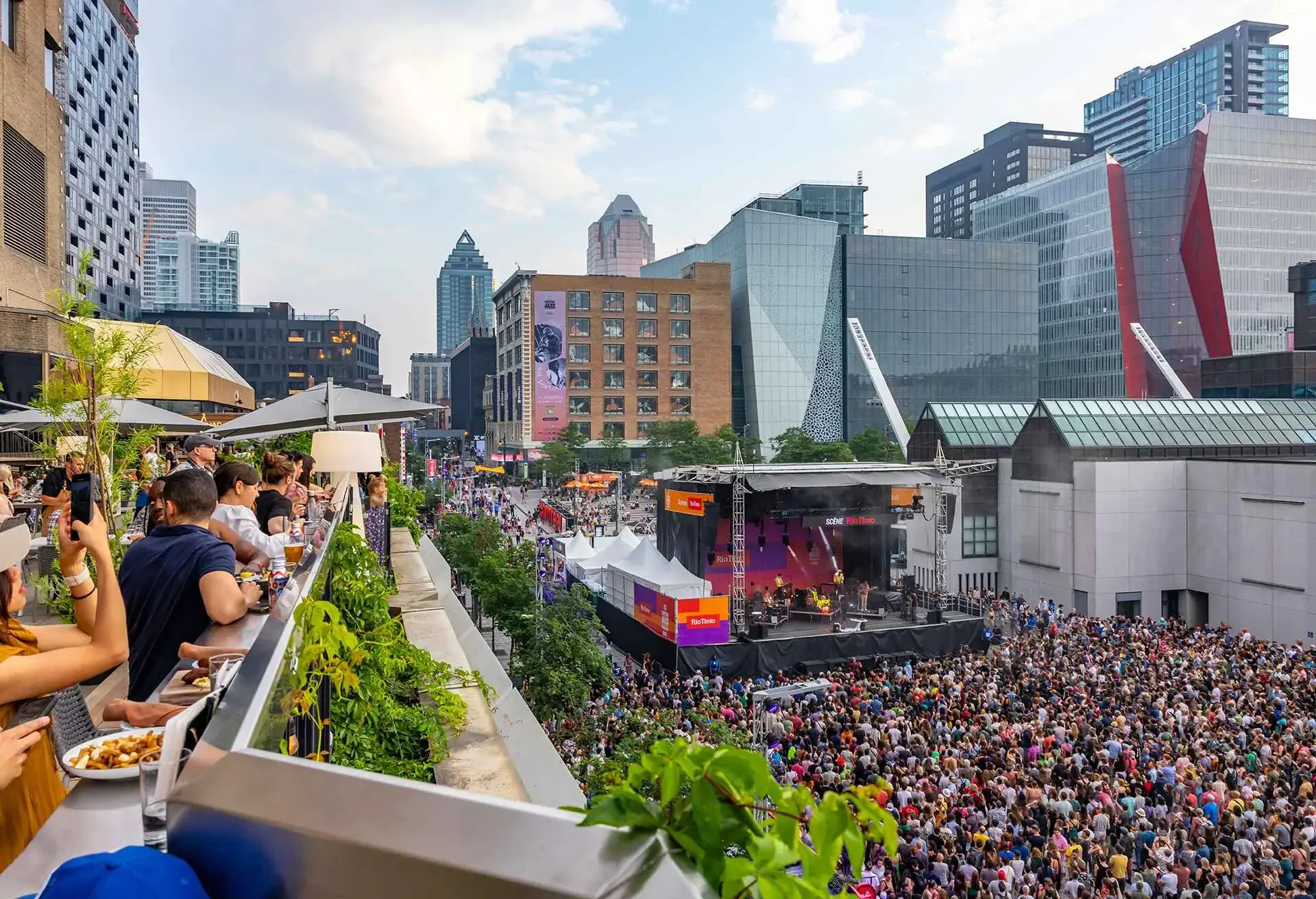 A large crowd attending an outdoor concert in a bustling city with modern buildings in the background