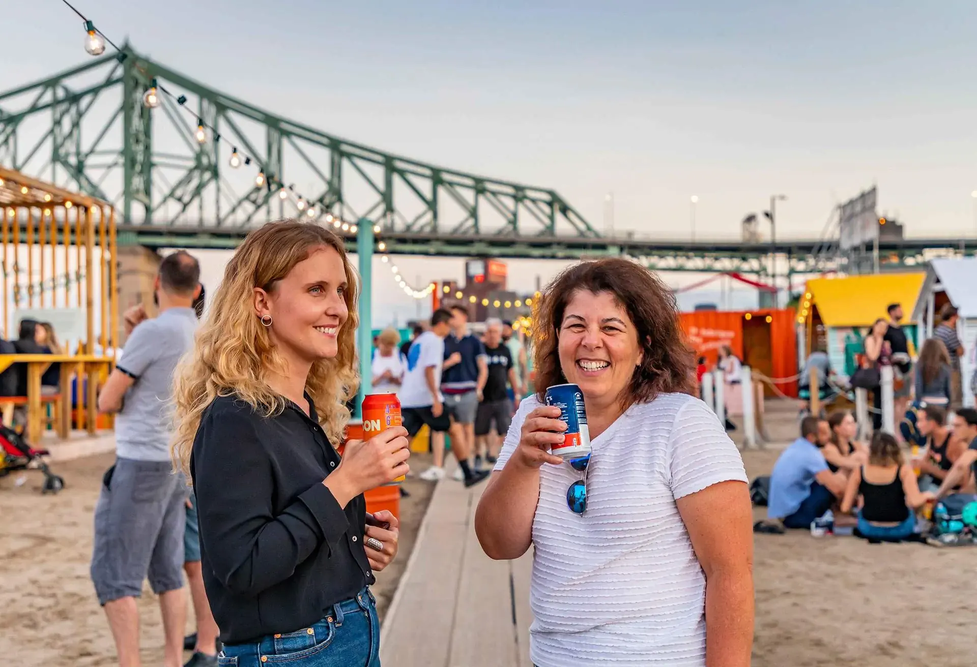 Two people drinking beverages at an outdoor festival near a bridge