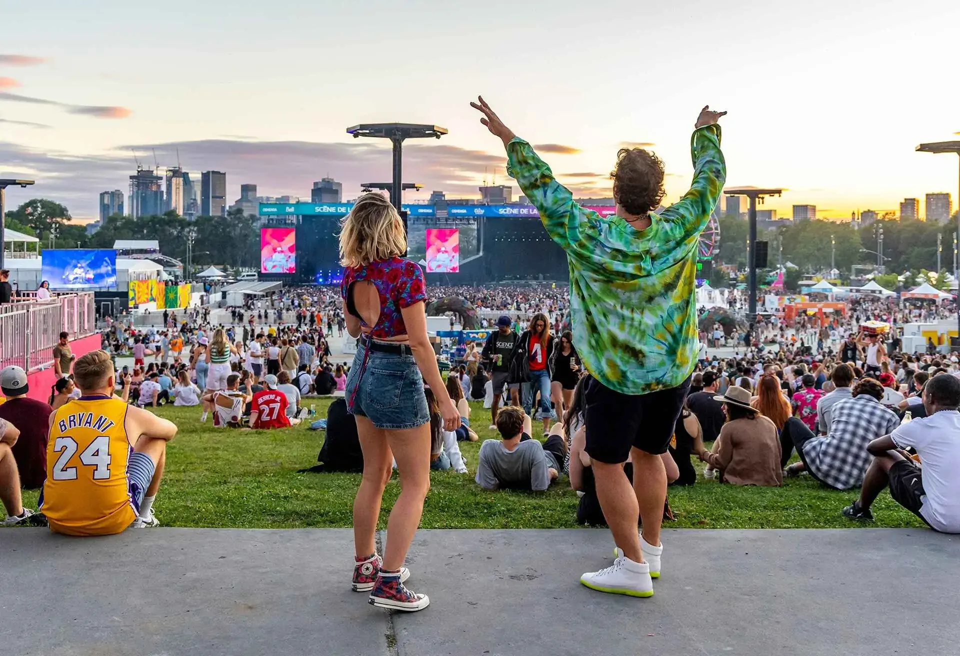 Two people enjoying a large outdoor concert with a crowd and stage in the background at sunset
