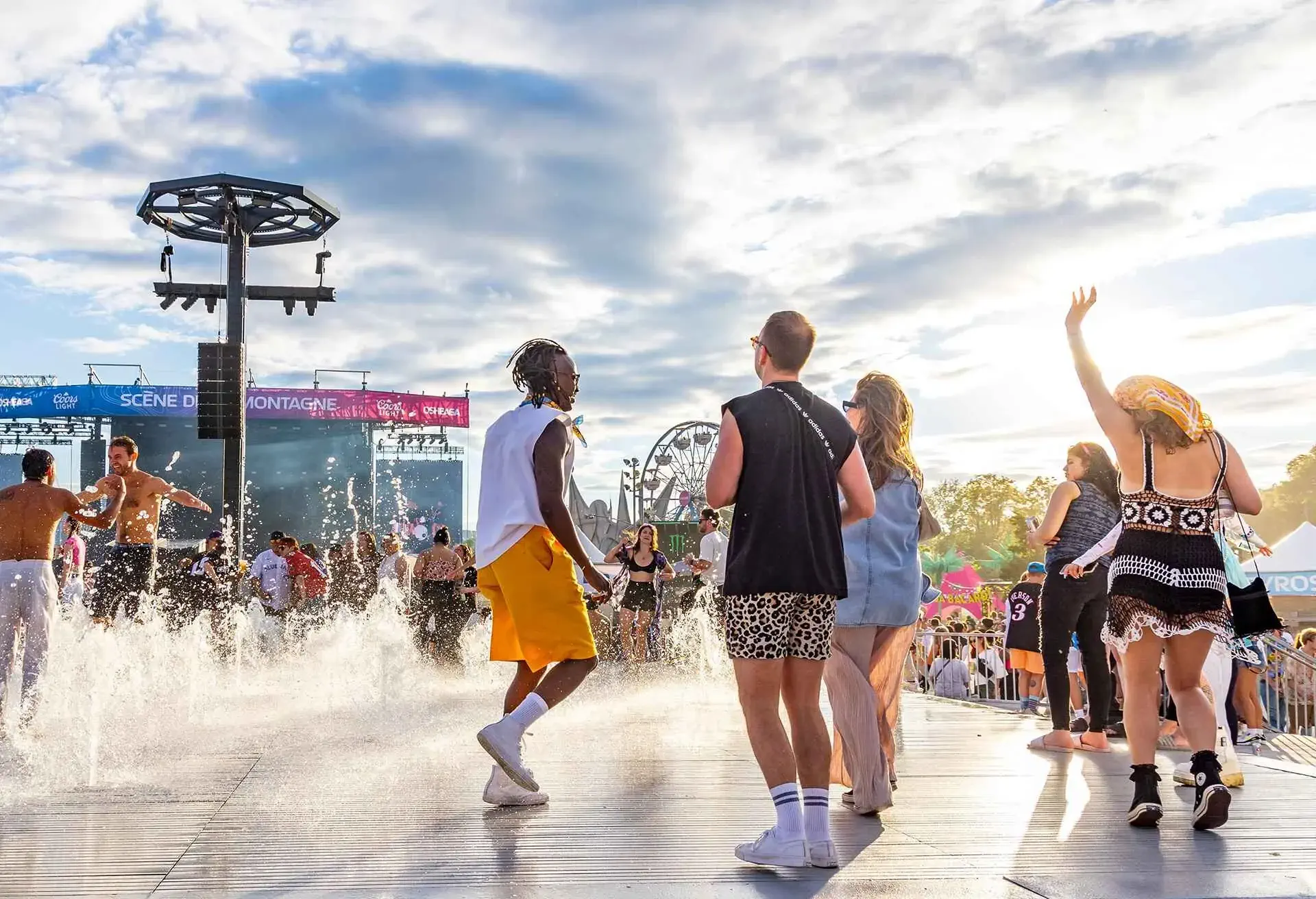 People enjoying a music festival with water features and Ferris wheel under a cloudy sky.