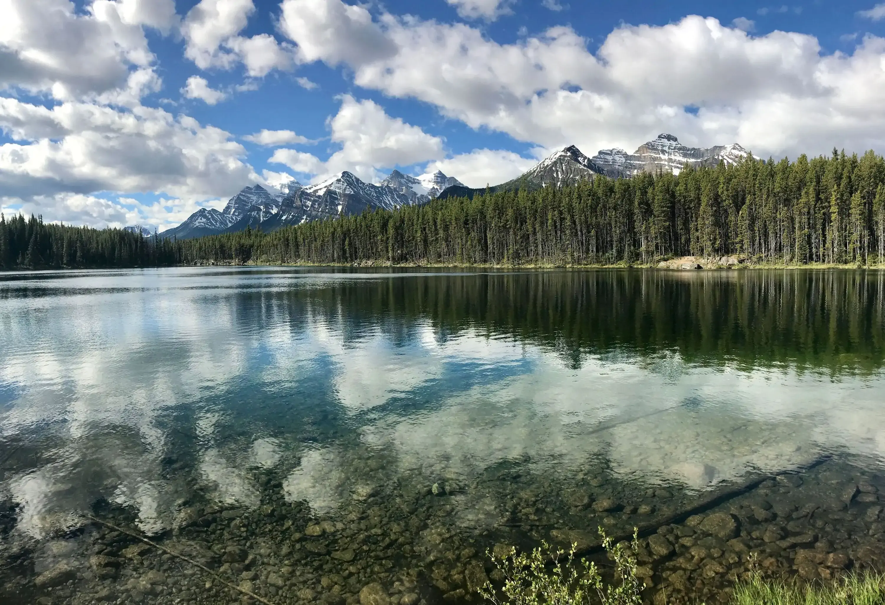 A clear water lake lined with tall green trees with a view of snow-dusted rocky mountains against the cloudy blue sky.