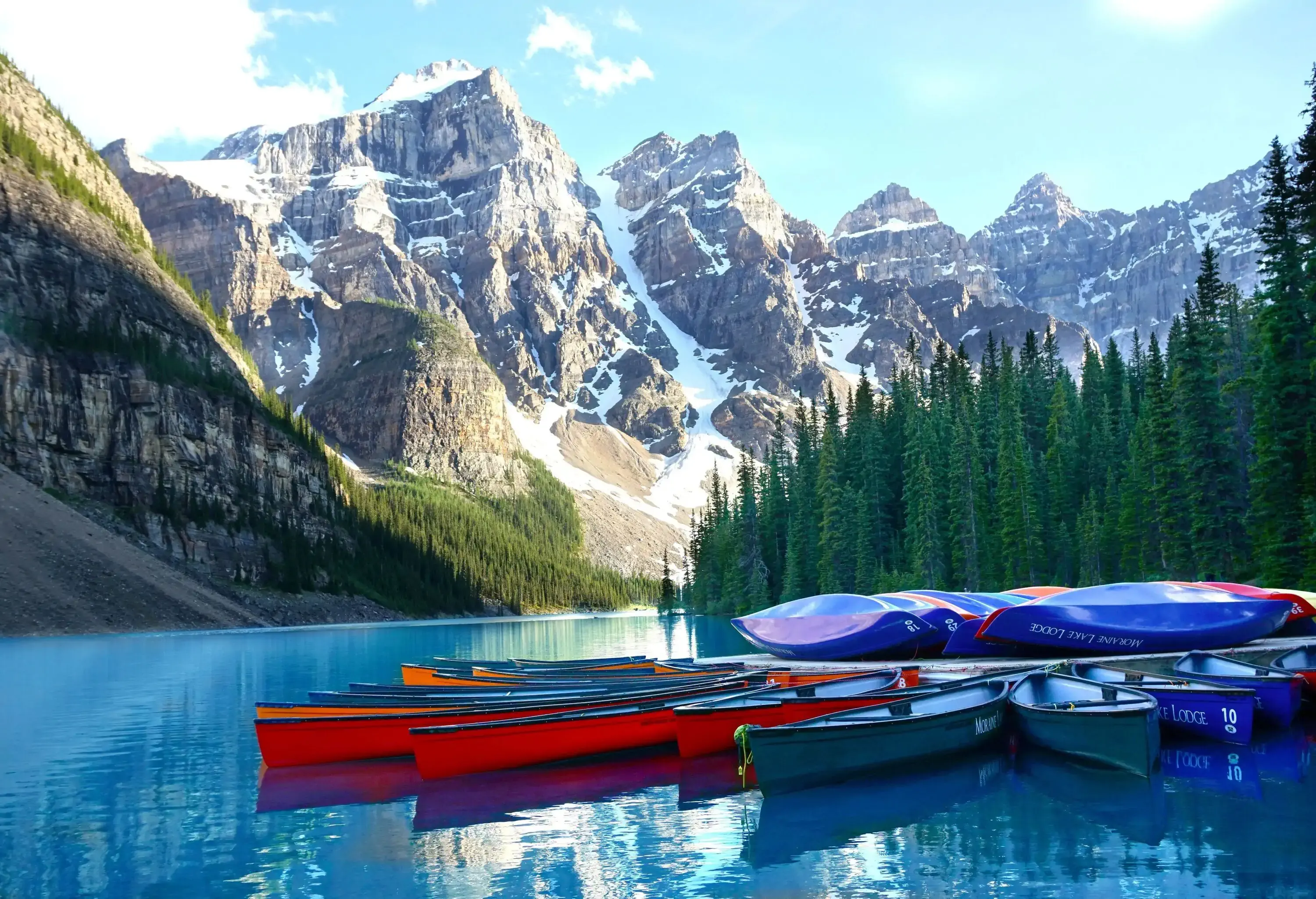 Colourful boats docked on a calm blue lake bordered by tall bushes and steep mountains covered with snow.
