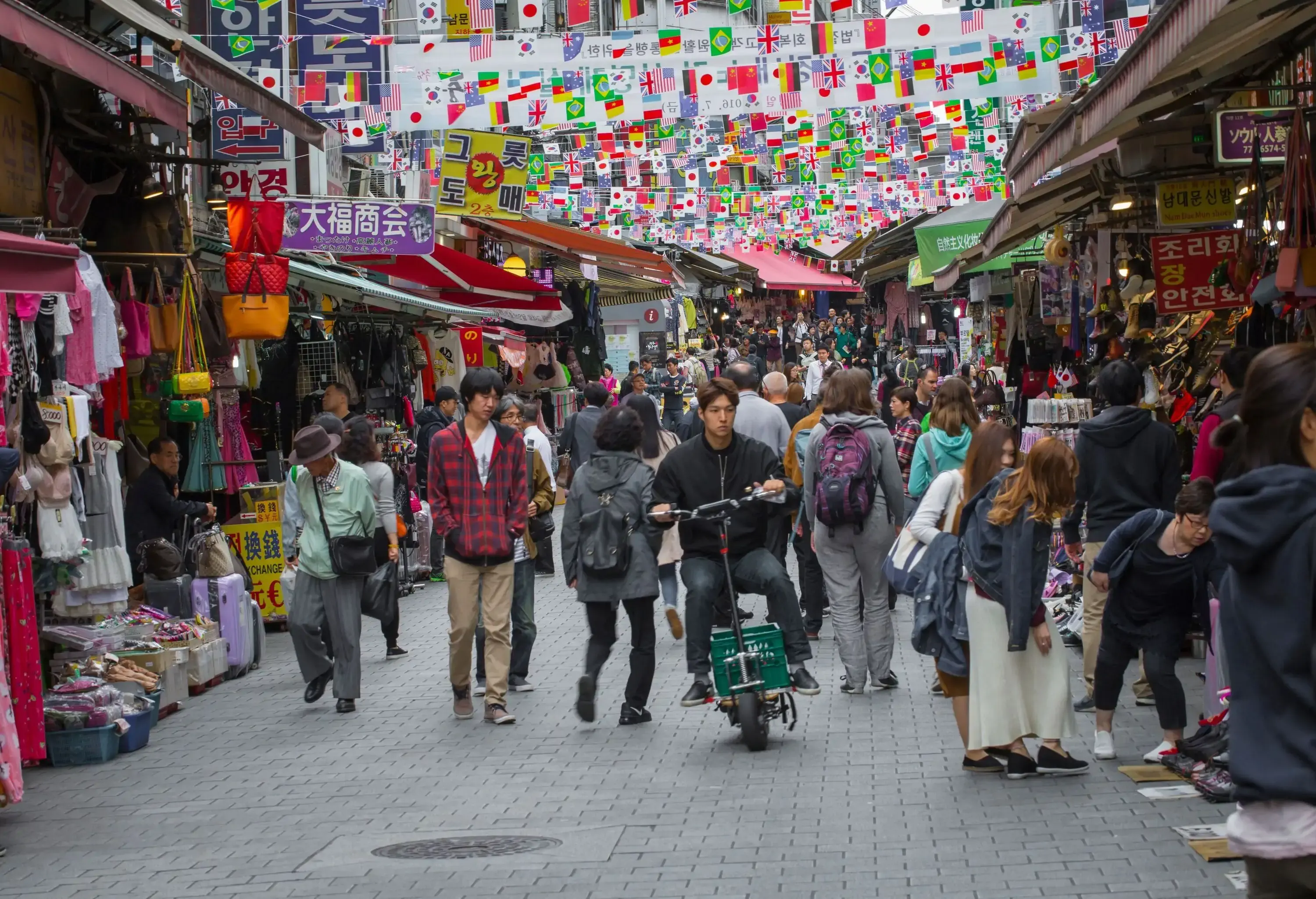 A crowded street lined with vendor stalls selling assorted souvenir items under hanging banners of country flags.
