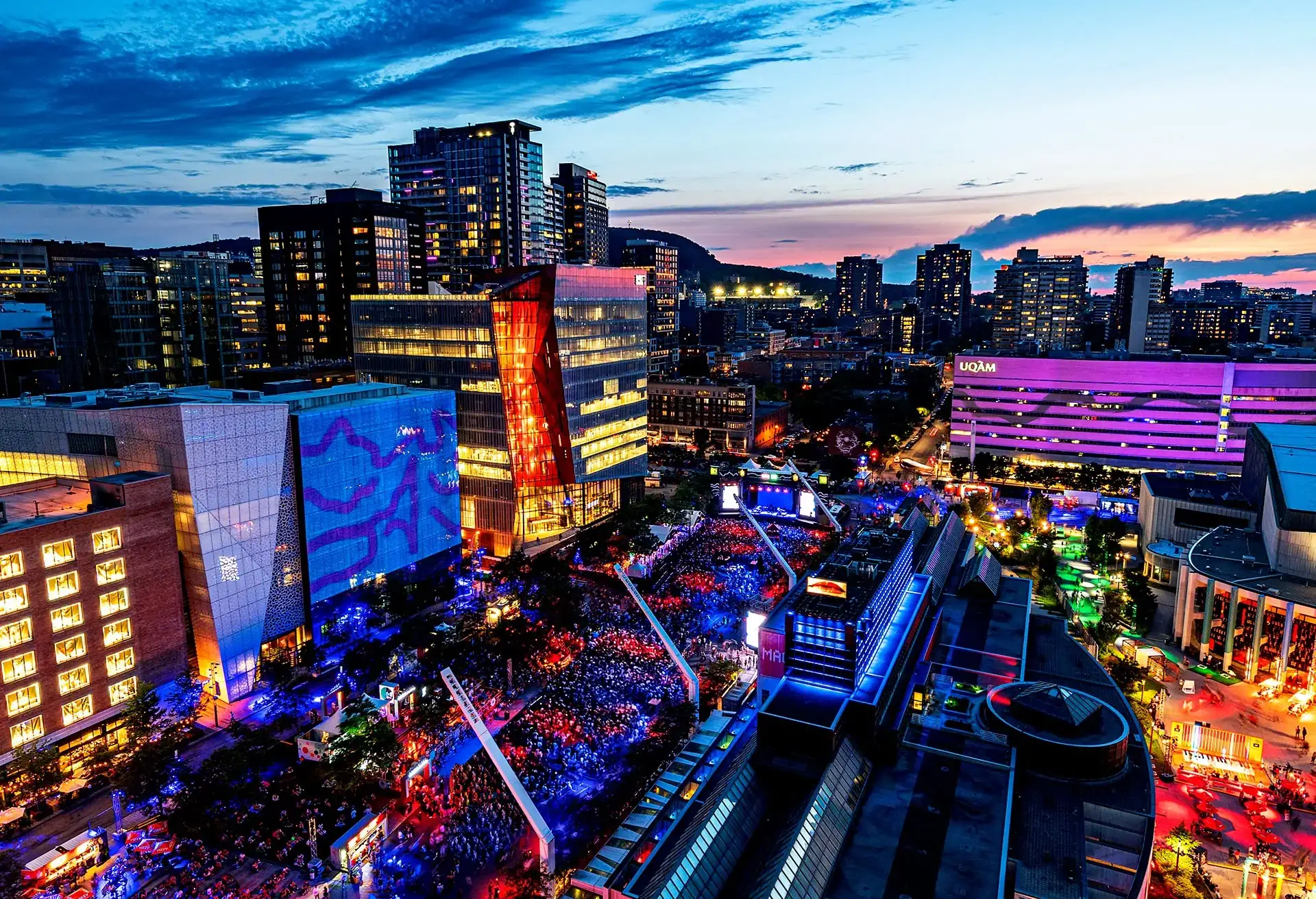 An aerial view of a vibrant outdoor music festival with colorful lights and modern buildings at dusk