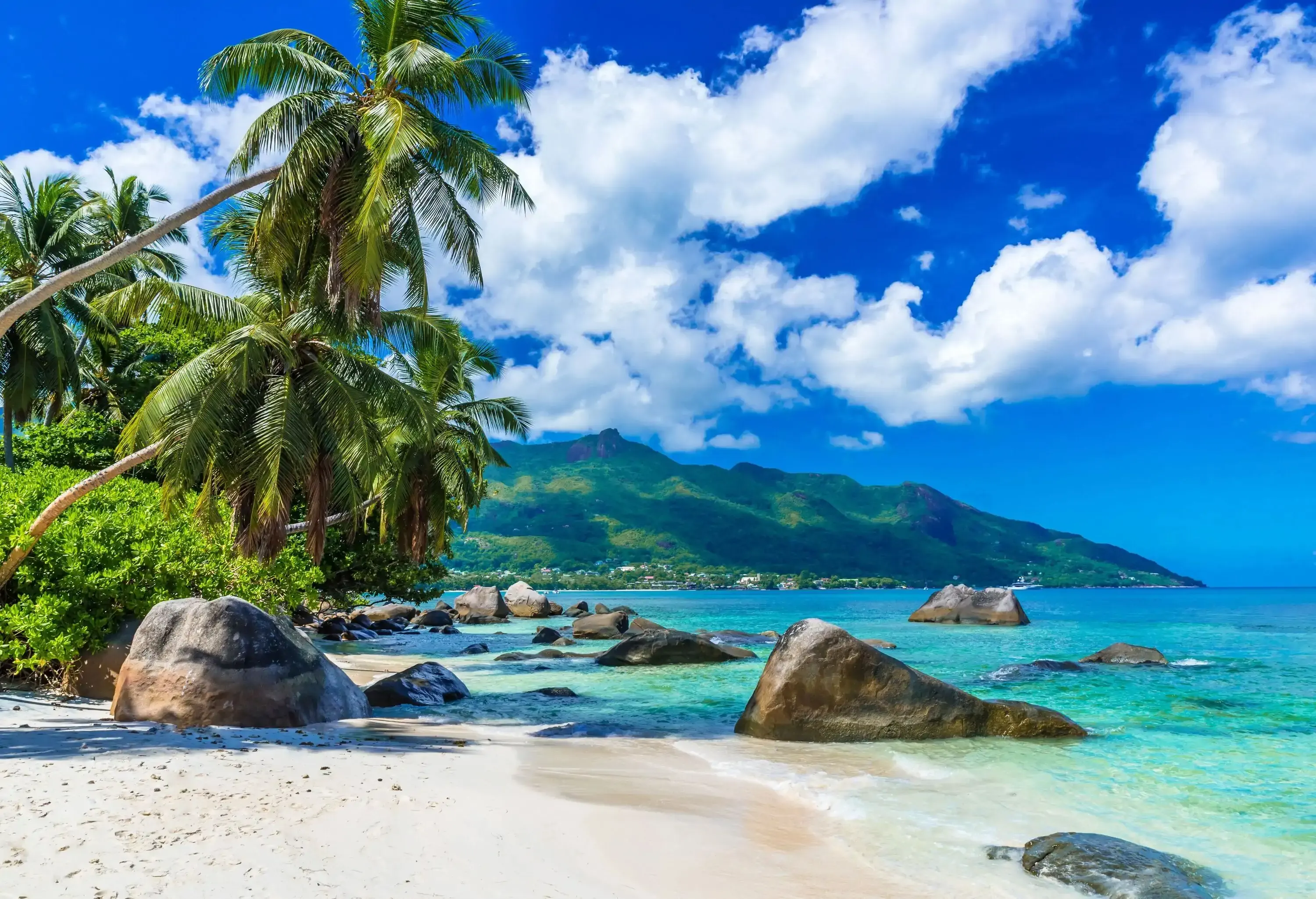 A tropical beach on a serene island, adorned with large rocks along the shore, with a backdrop of lush green mountains across the sparkling sea.