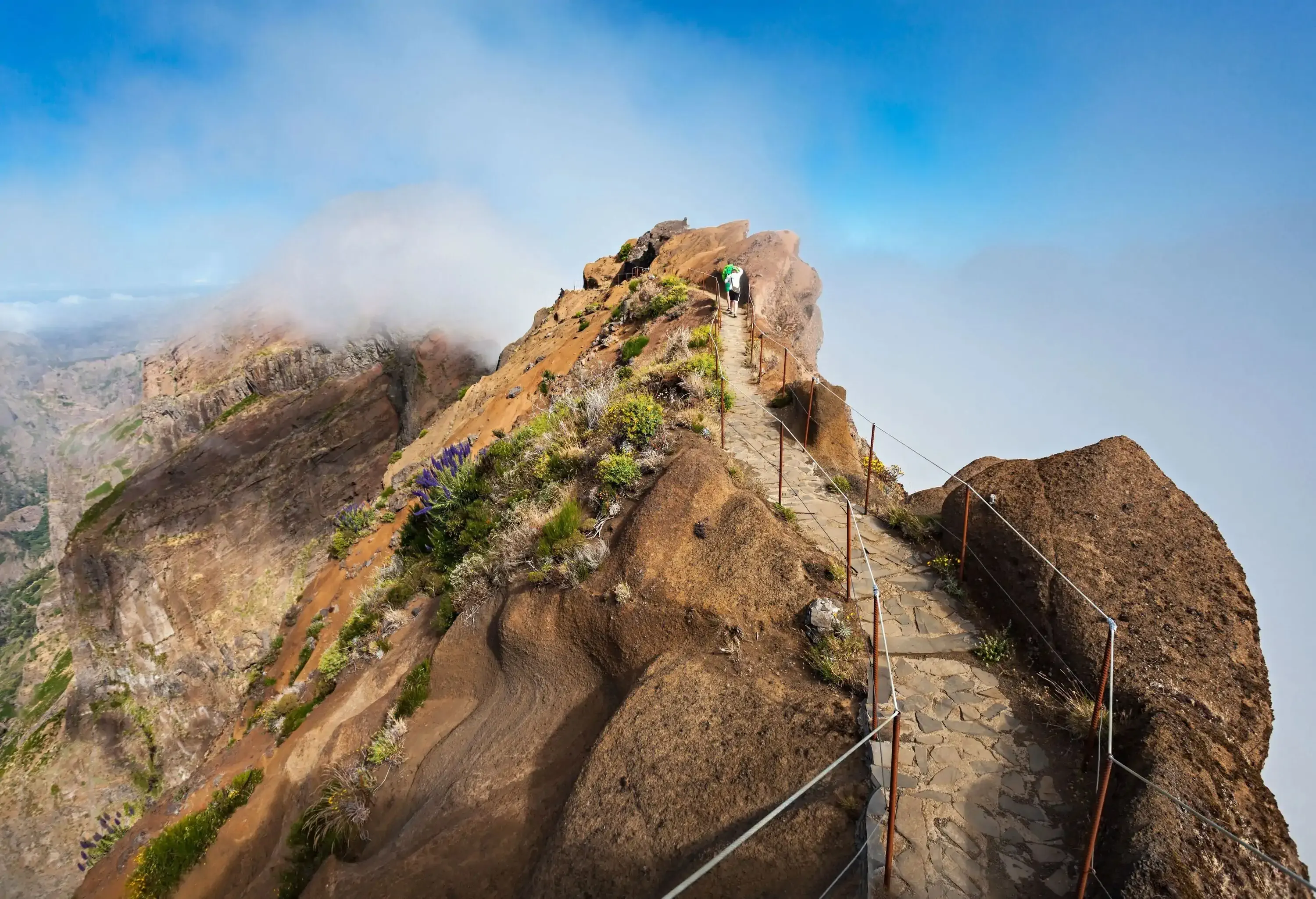 A person trekking on a stone path on top of a mountain ridge with views of misty valleys below.
