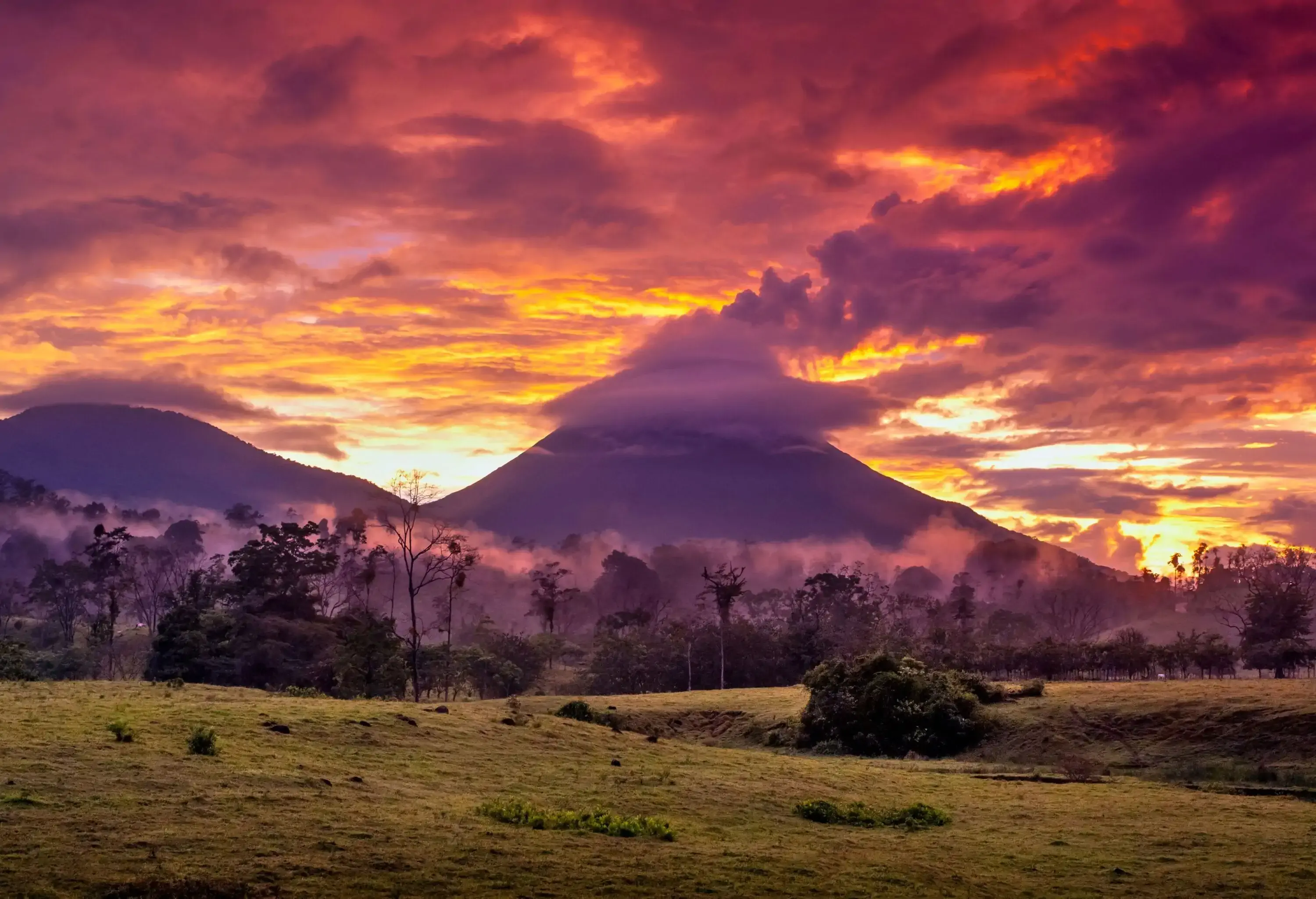 As the sun sets, a mystical ground fog envelopes the lush forest surrounding the Arenal Volcano, while a cloudy sky overhead casts a warm glow over the stunning landscape.