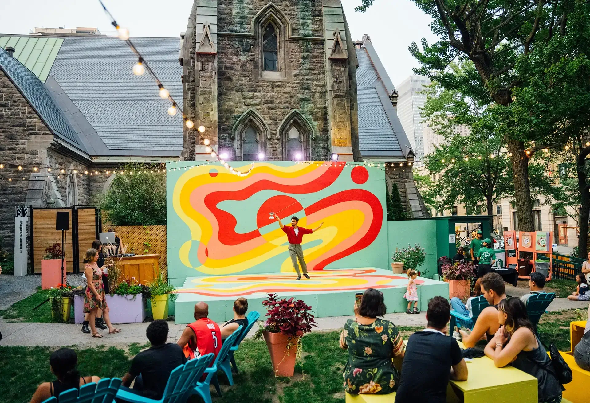 Performer in red shirt on colorful outdoor stage with audience seated and standing, in front of historic stone building under string lights