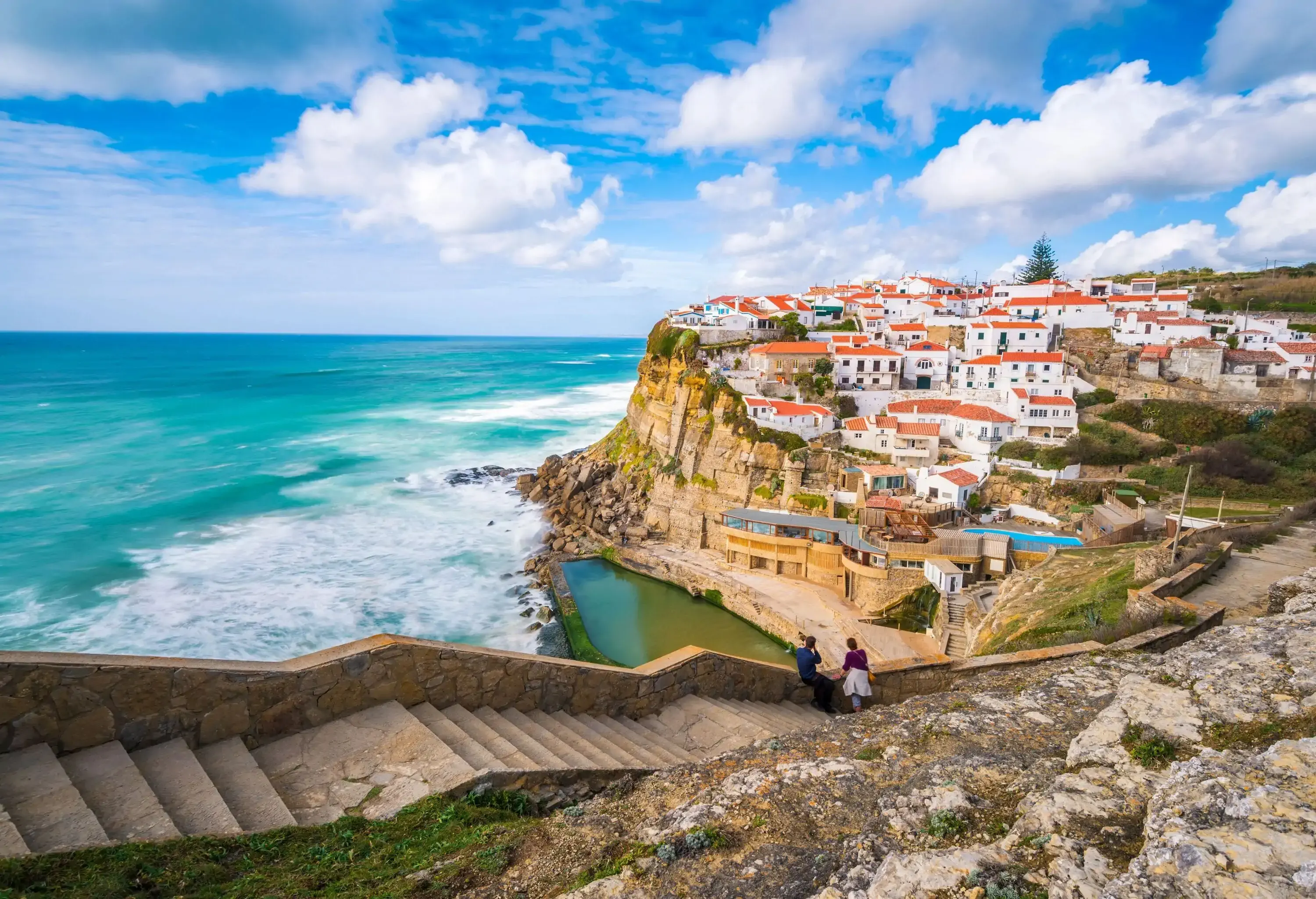 Two people are sitting on the steps leading to the coastal village, enjoying the scenic view.