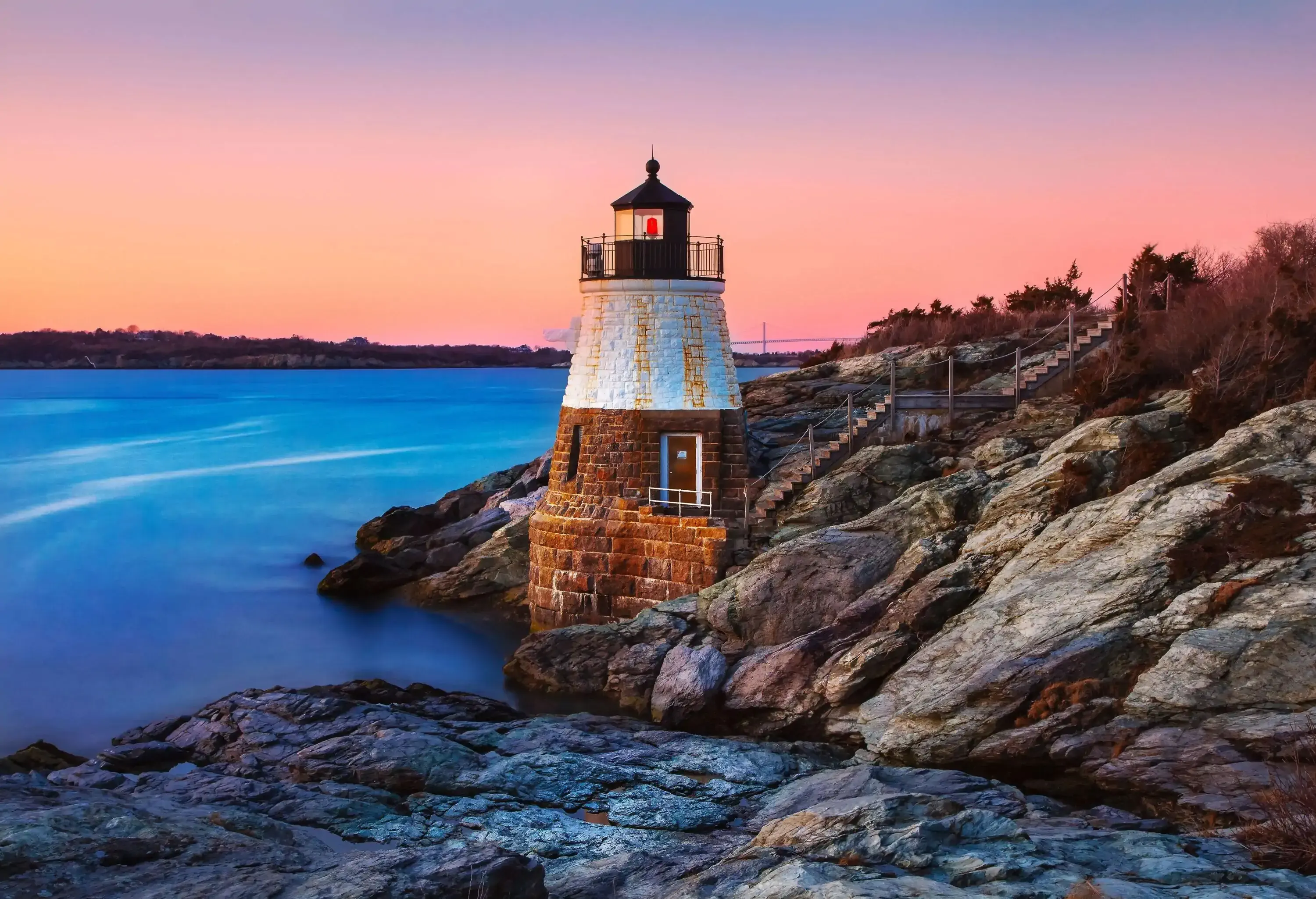 A lighthouse along a rocky shoreline, with faint illumination from the pink-hued sky. 