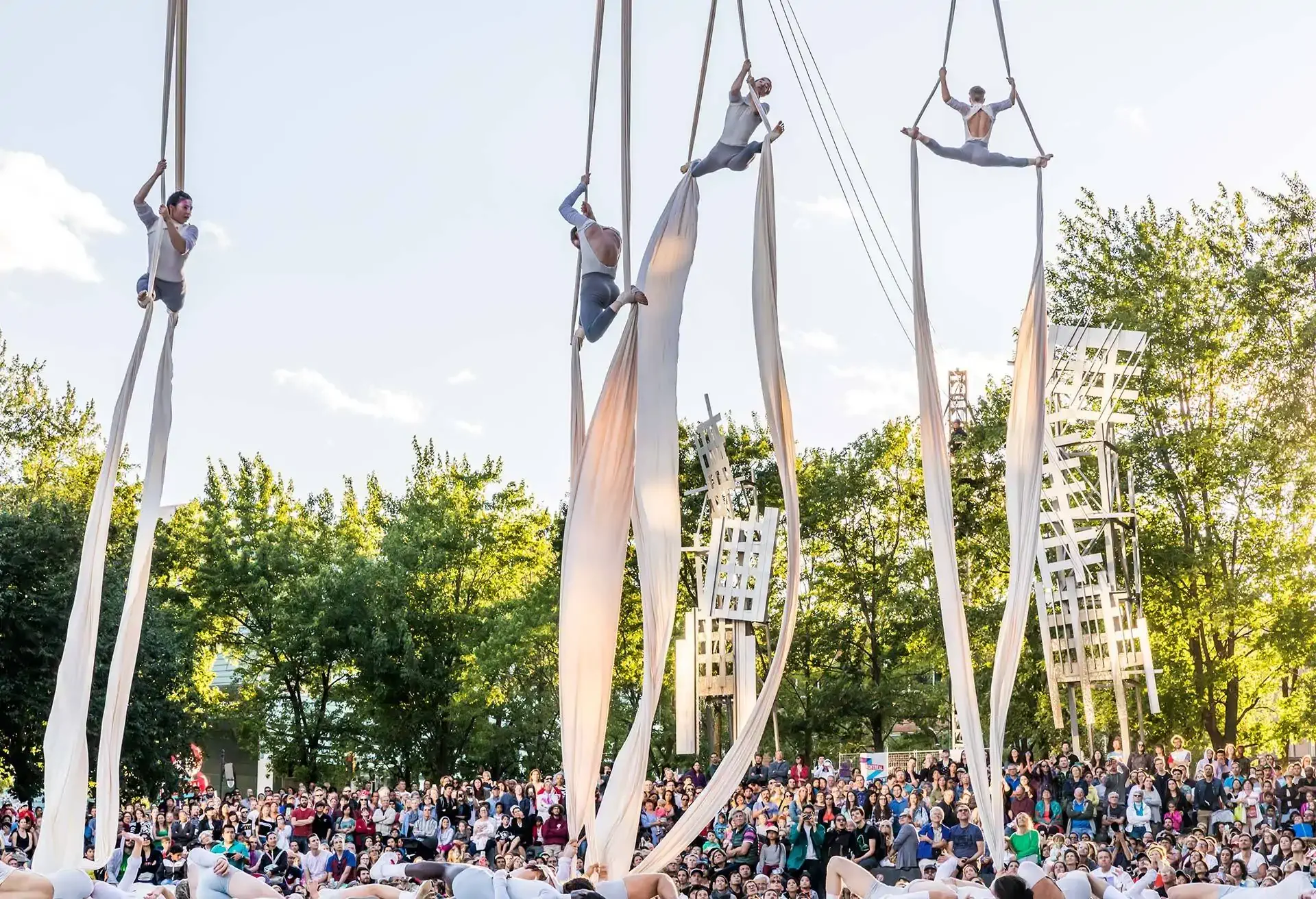 Aerial performers on ropes in front of a large outdoor audience and trees.