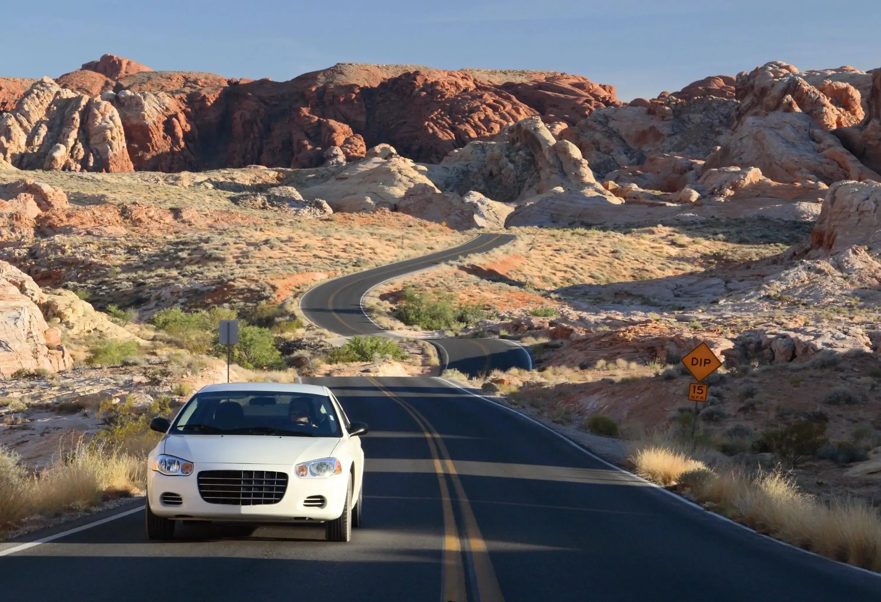A white car drives away from the winding road, leaving behind the vibrant landscapes of Valley of Fire State Park, with majestic mountains standing tall in the distance.