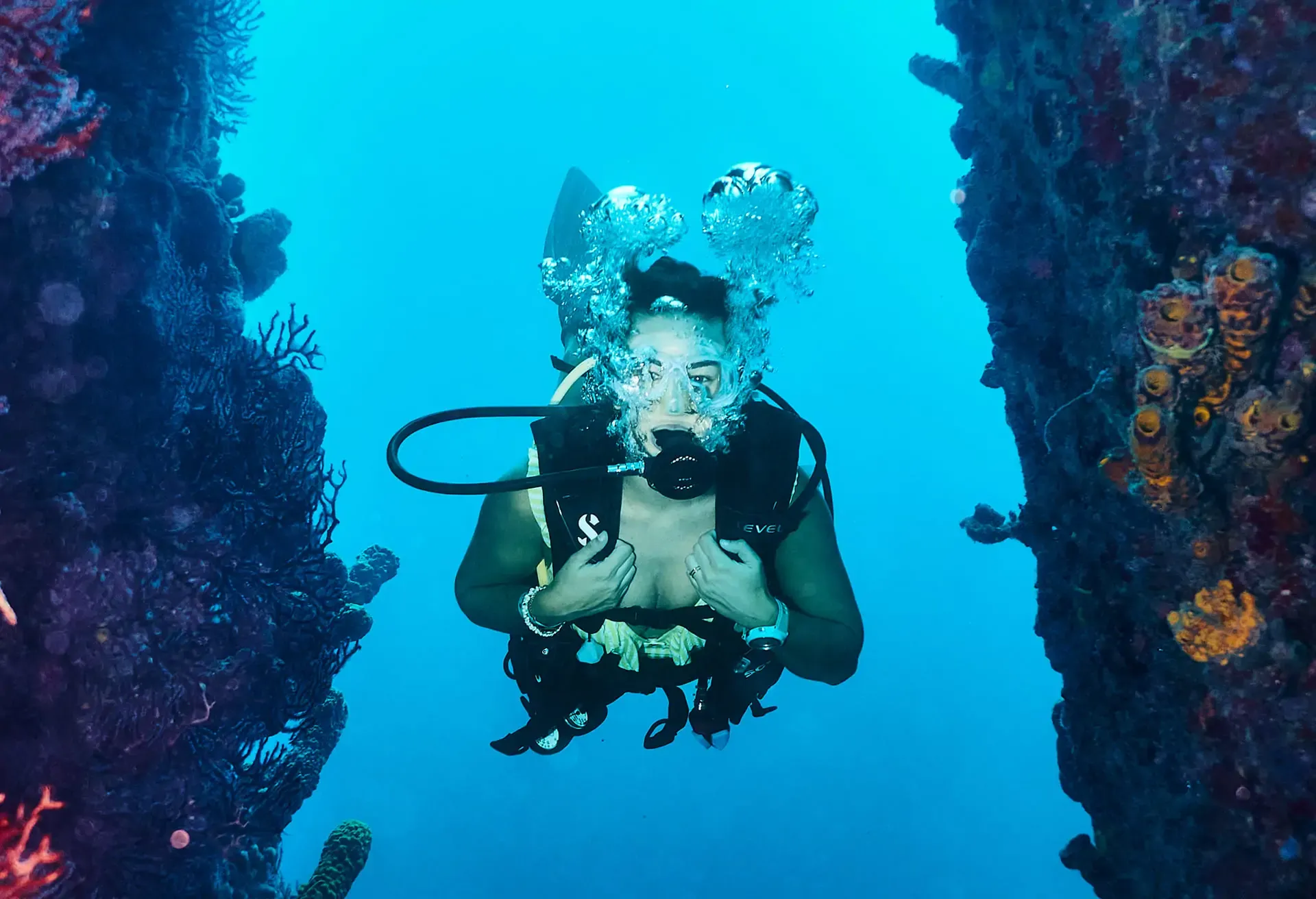 A scuba diver swimming through a narrow coral reef opening in deep blue water.