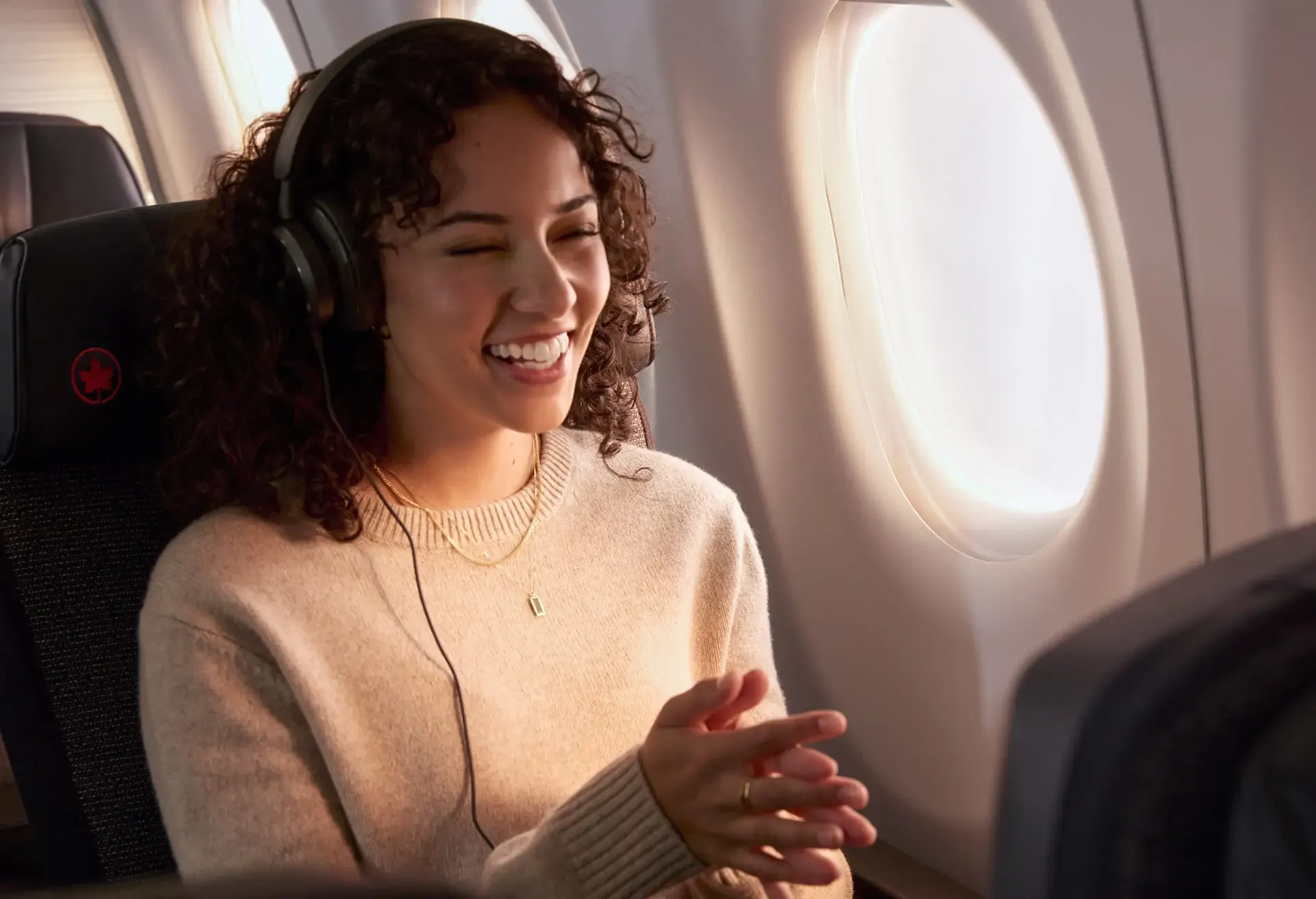 A cheerful woman with curly hair wearing headphones and sitting by an airplane window.